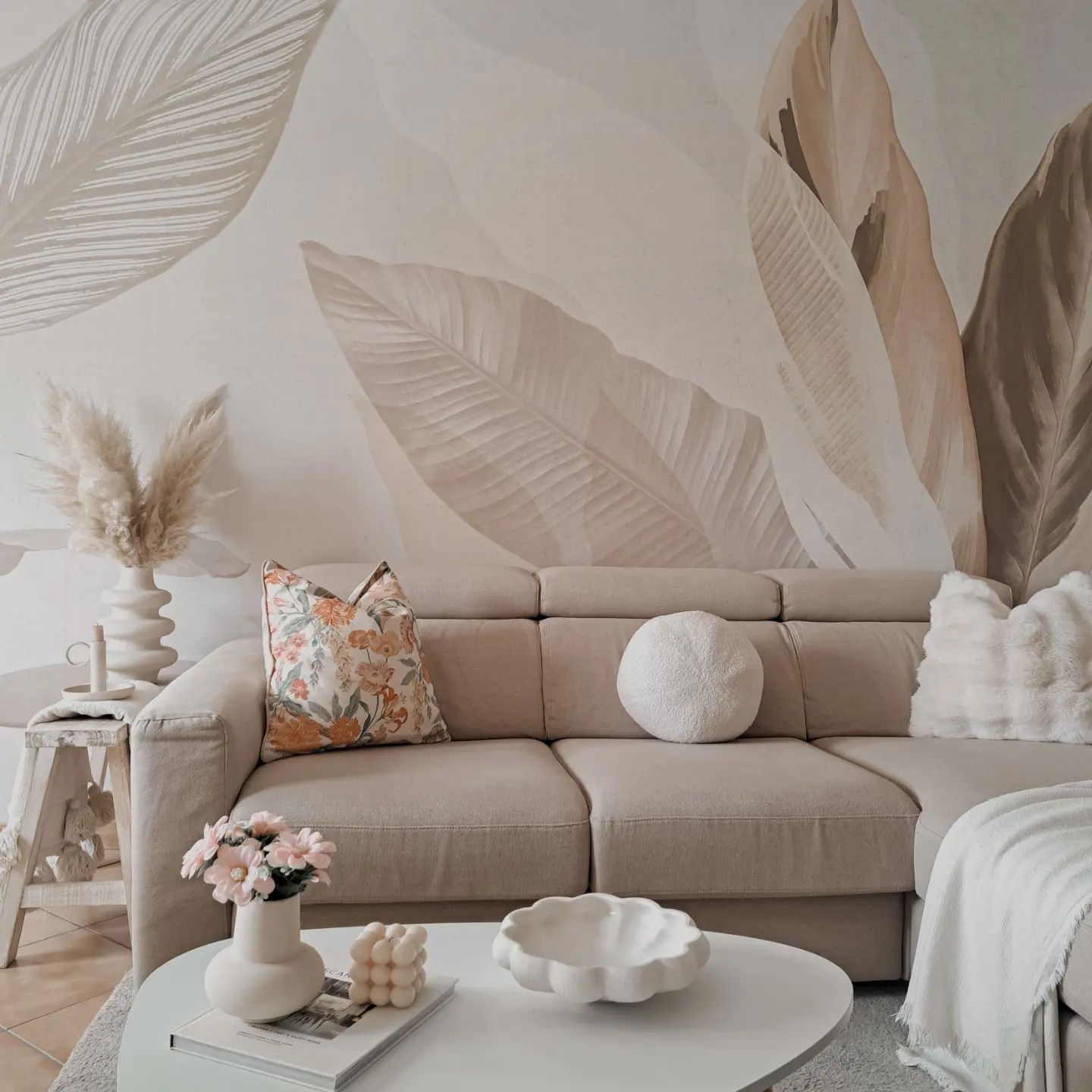 Nøytral stue med beige seksjonssofa, blomstrete puter og blad-mønstret tapet.
