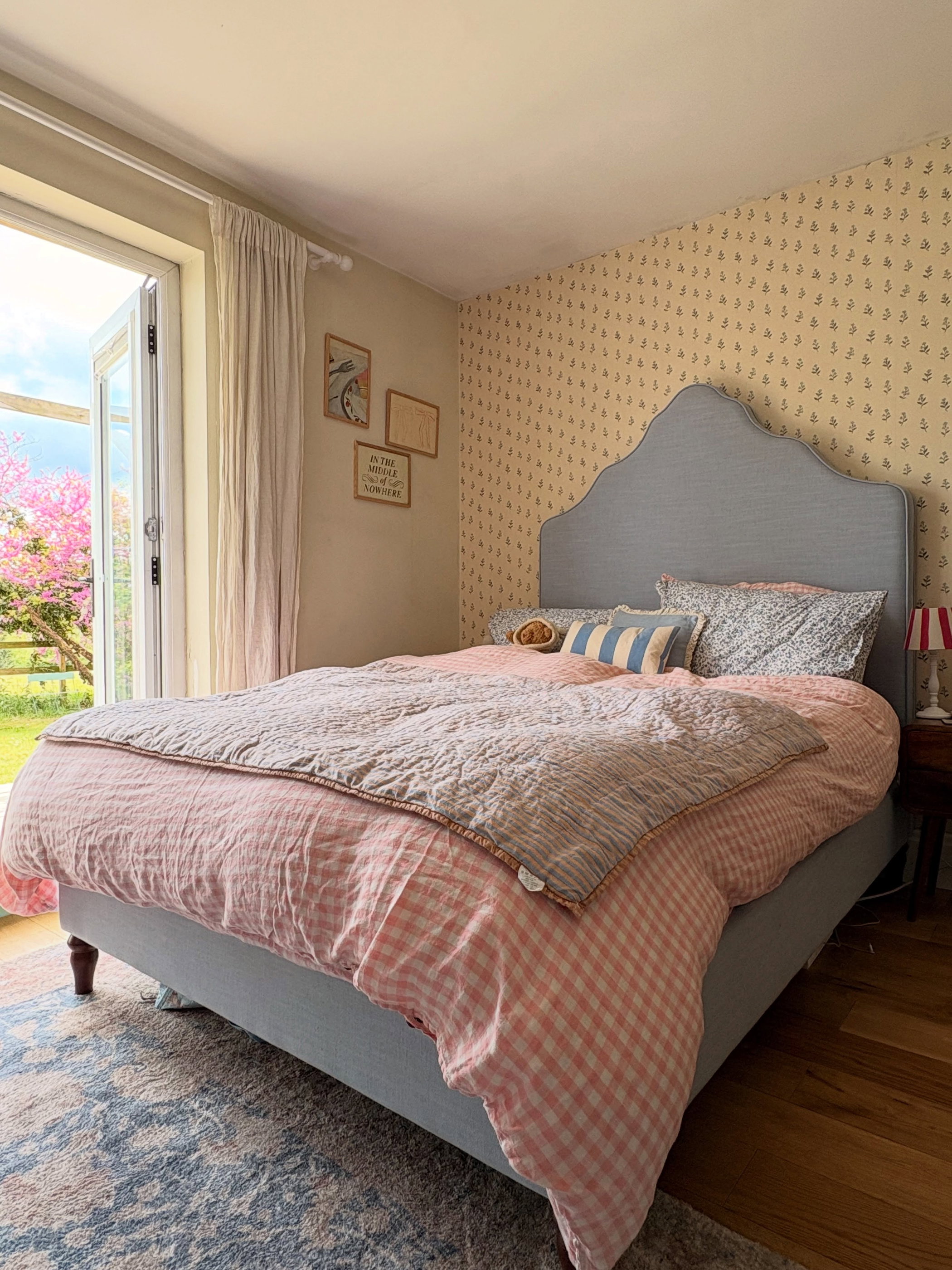 Cozy bedroom with a pink gingham duvet, striped quilt, and light blue headboard. Open doors reveal a flowering tree.
