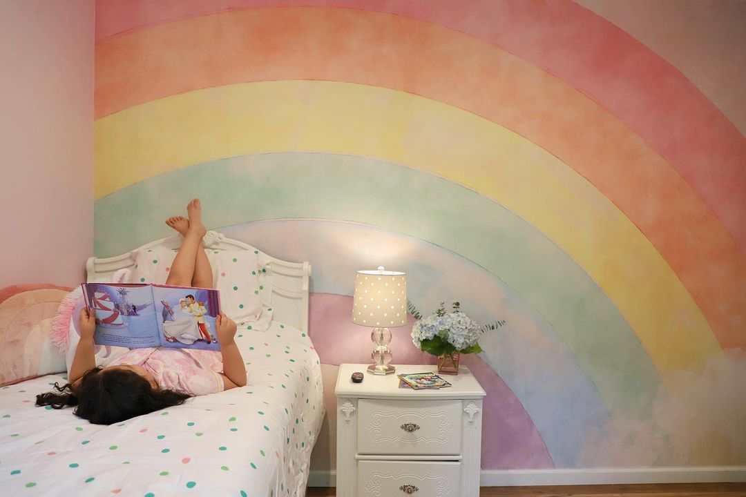 Mädchen liest im Bett mit Regenbogen-Wandbild. Nachttisch mit Lampe und Blumen.