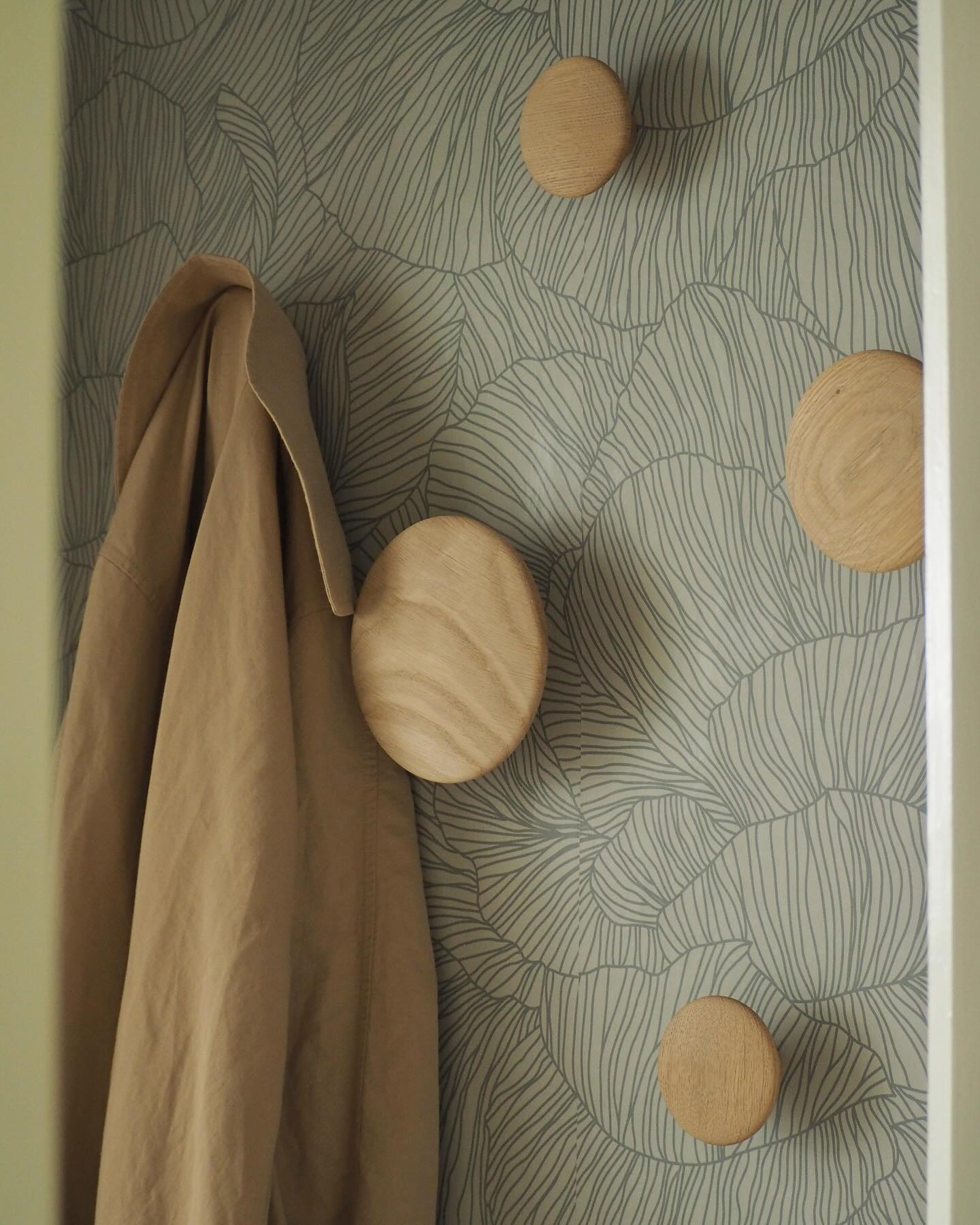 Cappotto color tan appeso a un gancio da parete in legno su carta da parati decorata.