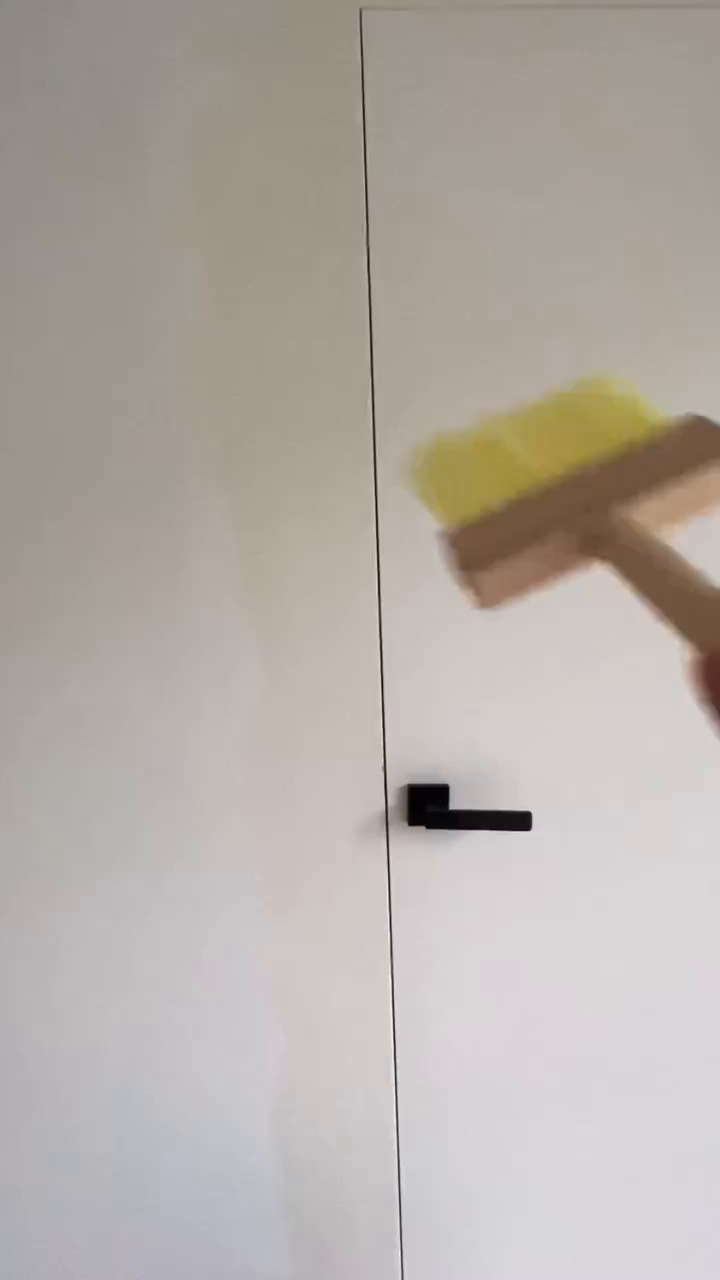 A hand holds a yellow-bristled brush near a white door with a black handle.