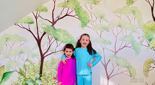 Dos chicas sonrientes en pijamas brillantes posan frente a un caprichoso mural de un árbol.