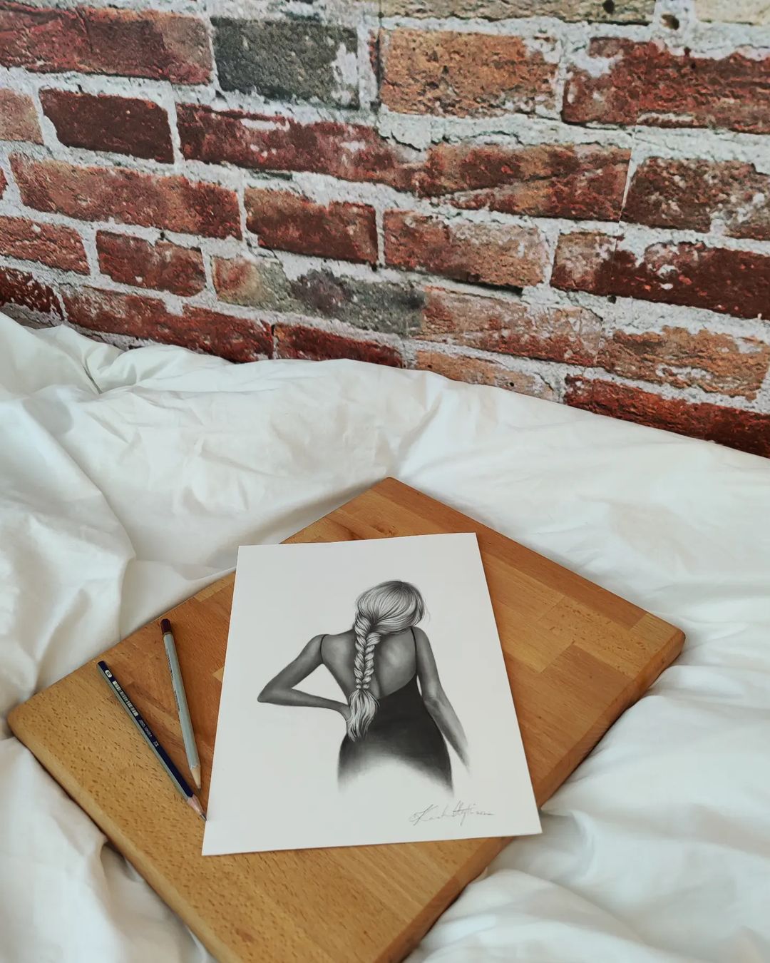 Drawing of a woman with a braid on a wooden board with pencils, on a white bed.