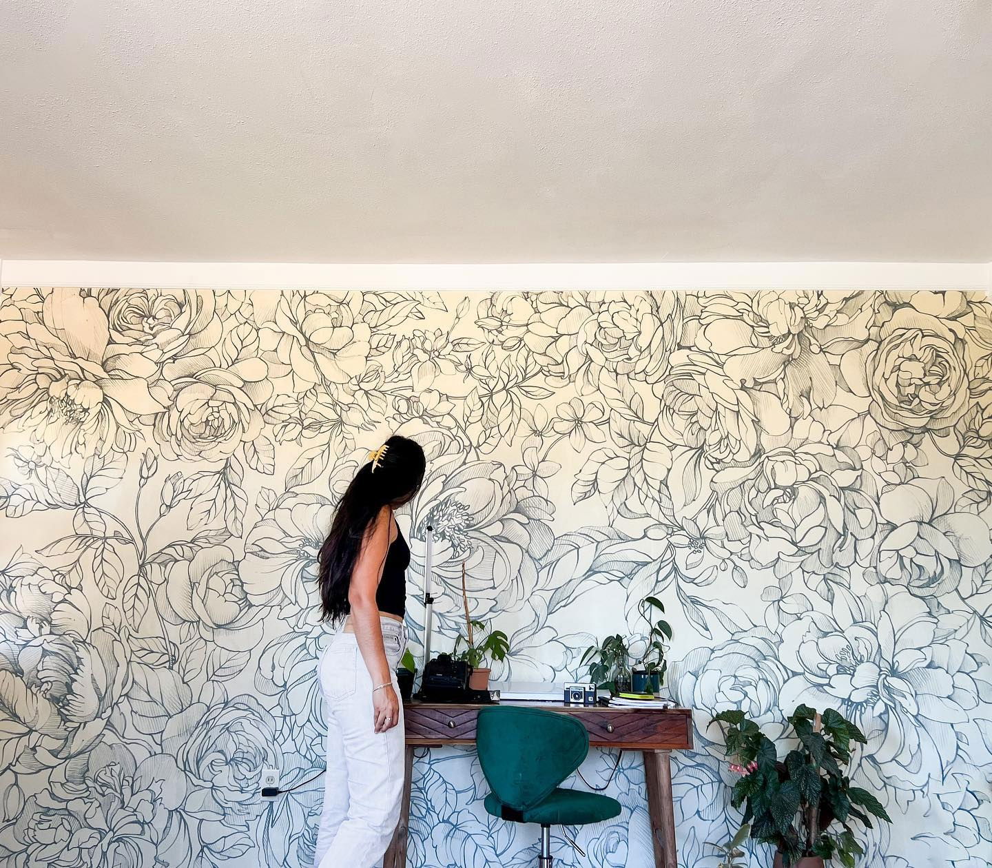 A woman stands in front of a floral wallpaper, next to a desk and green chair.