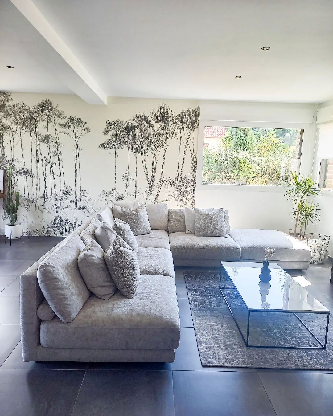 Wohnzimmer mit grauem Ecksofa, Couchtisch aus Glas und Baumwandbild.