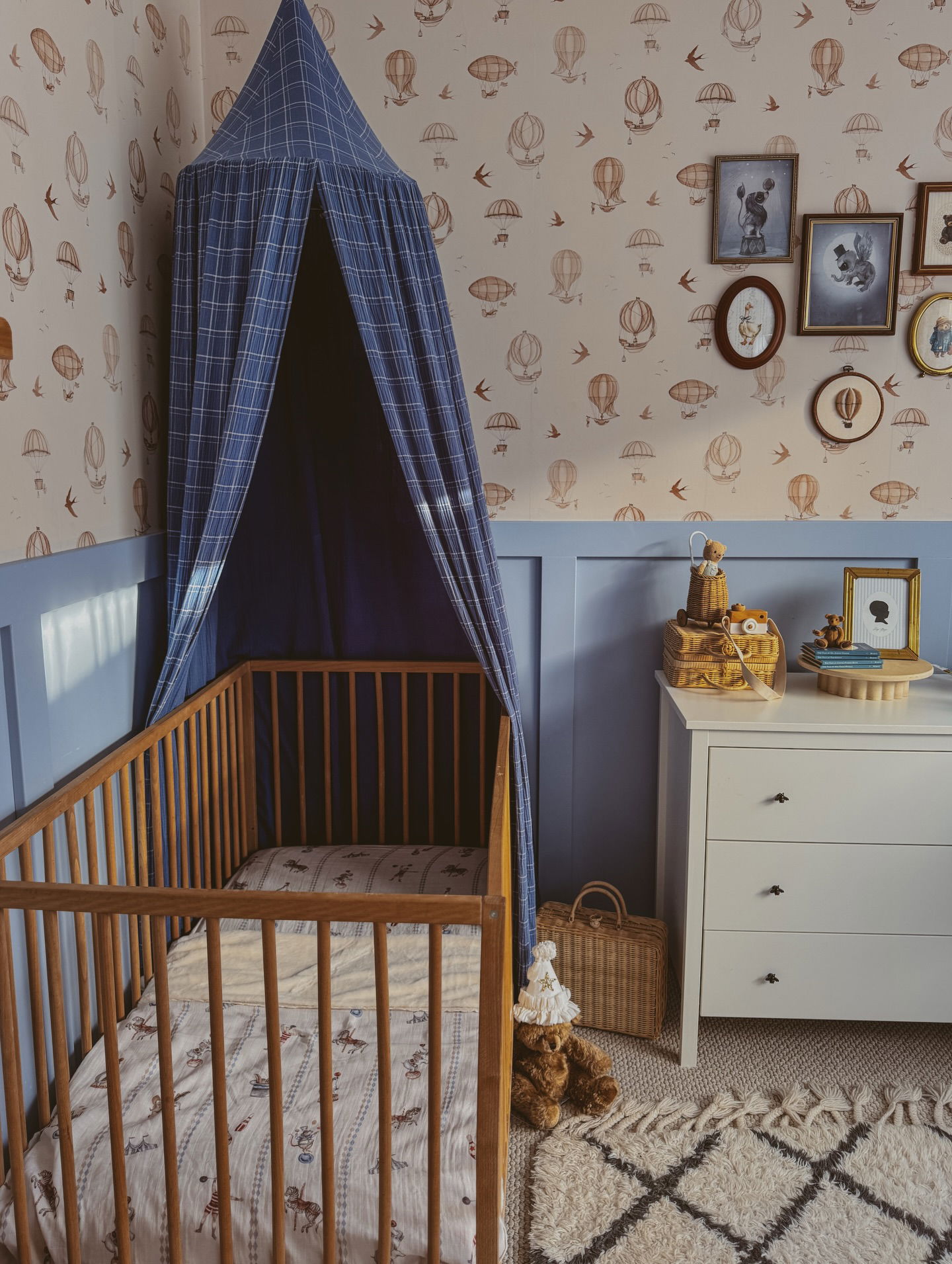 Chambre d'enfant avec berceau en bois, baldaquin à carreaux bleus, papier peint montgolfière et commode blanche.