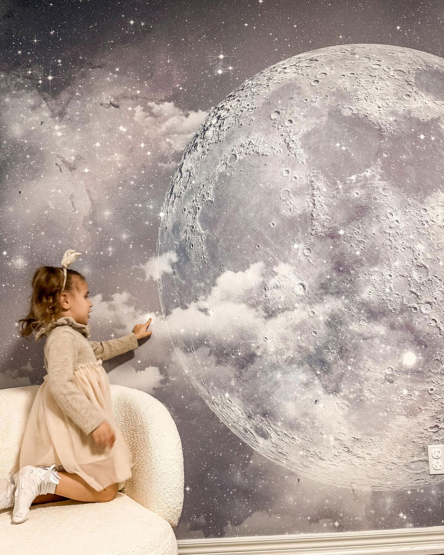 A young girl touches a moon and stars wallpaper. She wears a dress and sits on a white couch.