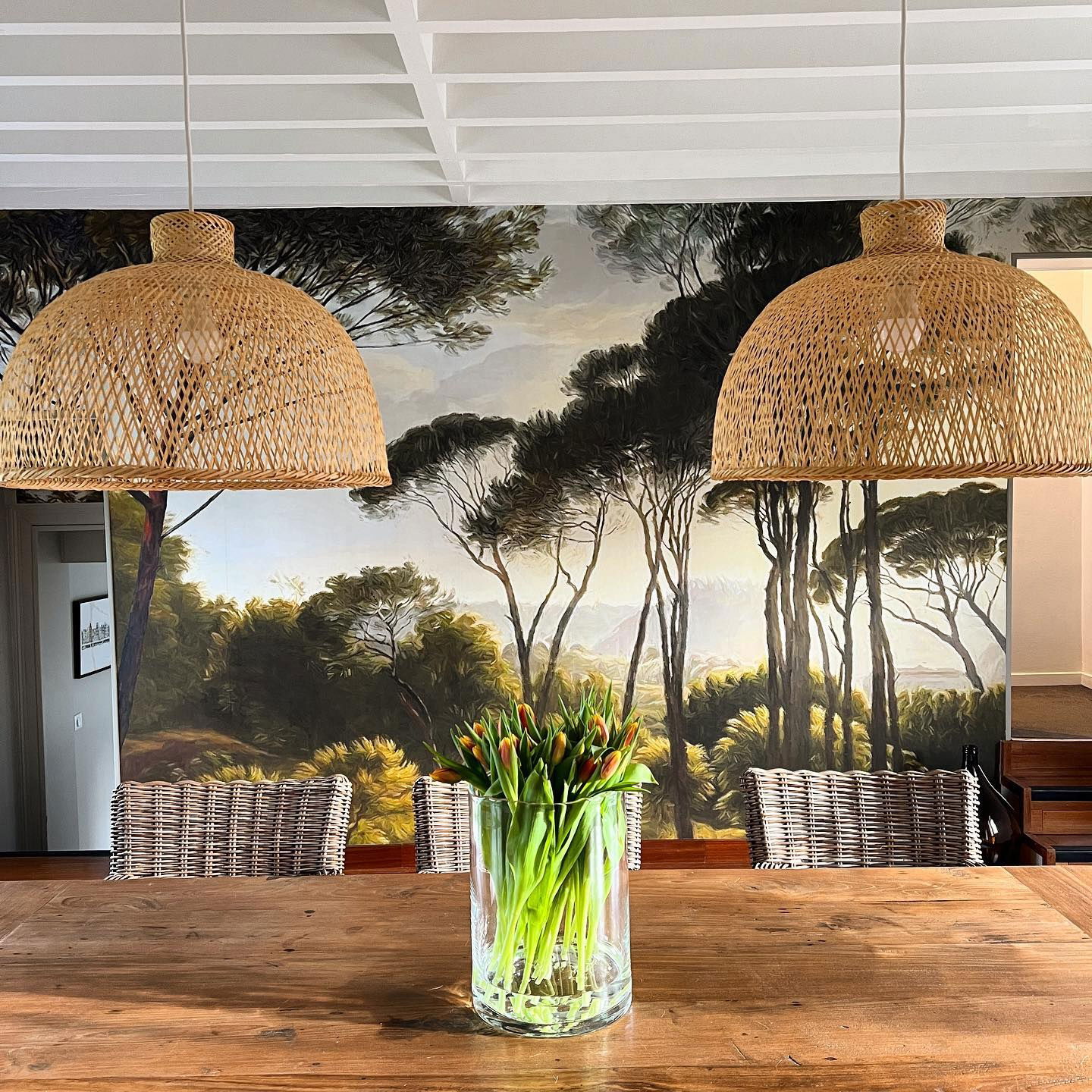 Dining room with wood table, woven chairs, tulip bouquet, and woven pendant lights against a forest mural.