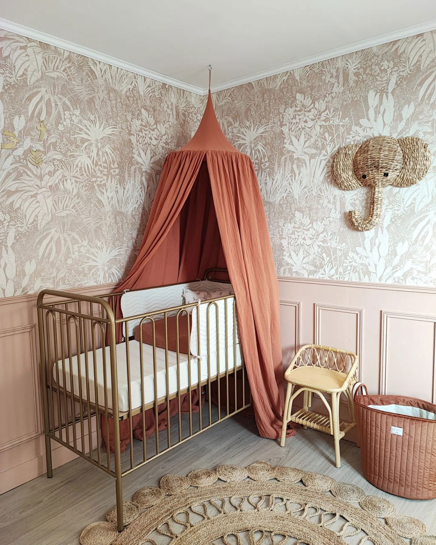 Habitación infantil con cuna dorada bajo dosel de coral, papel pintado de la jungla y cabeza de elefante tejida.