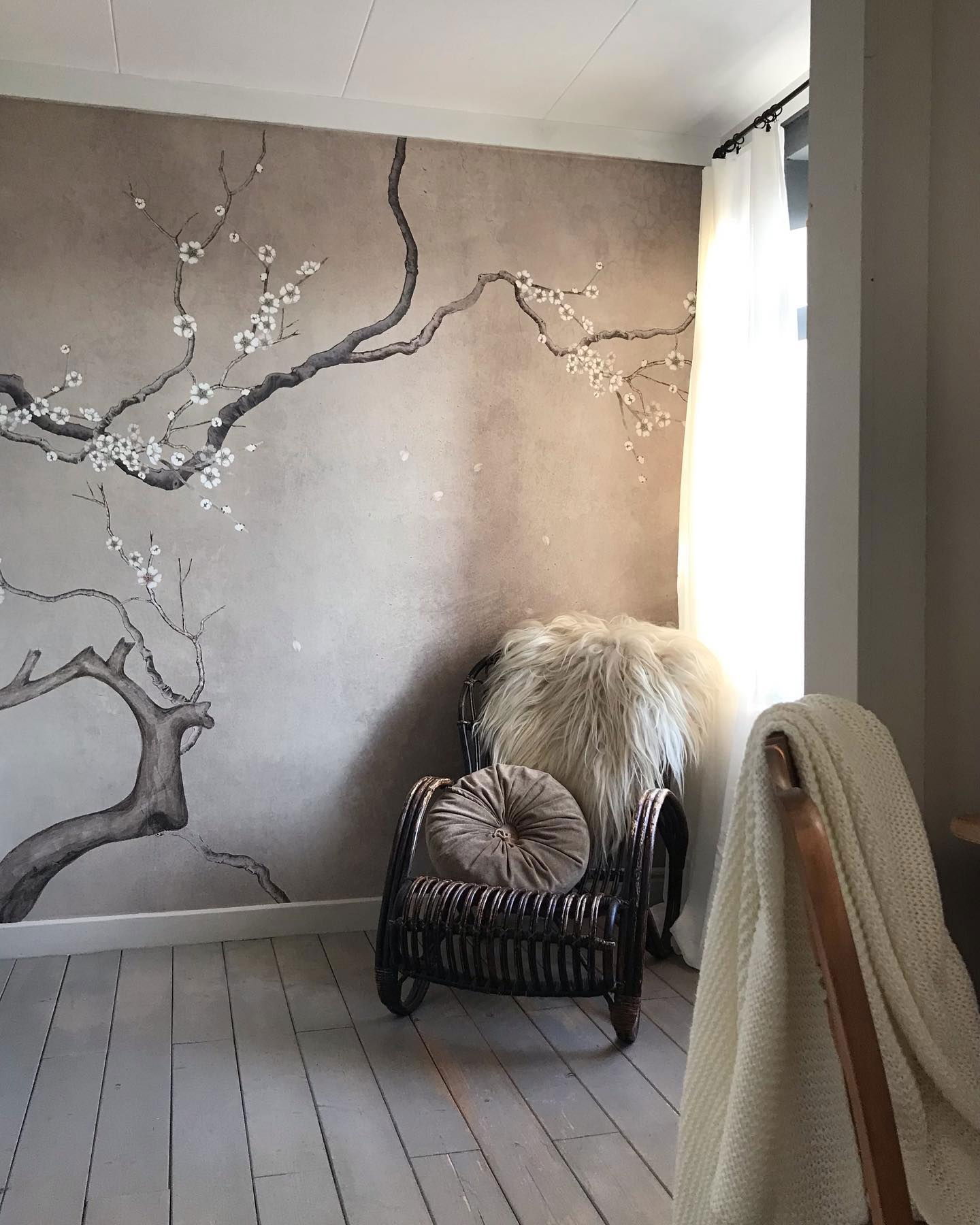 Hyggeligt hjørne af et værelse med blomstermaleri, kurvestol og bløde puder. Neutral farvepalet.