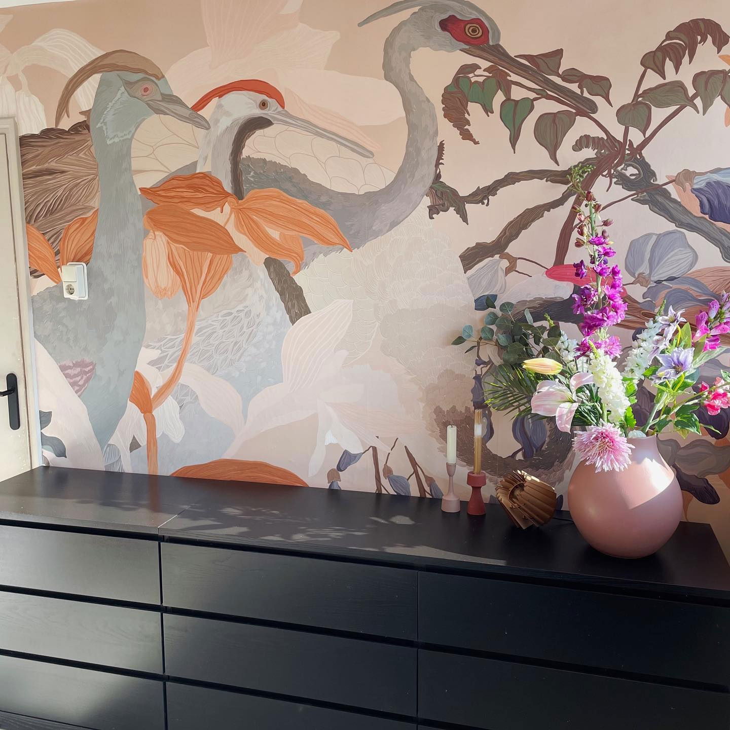 Interior with black dresser, floral mural, and pink vase with colorful flowers.
