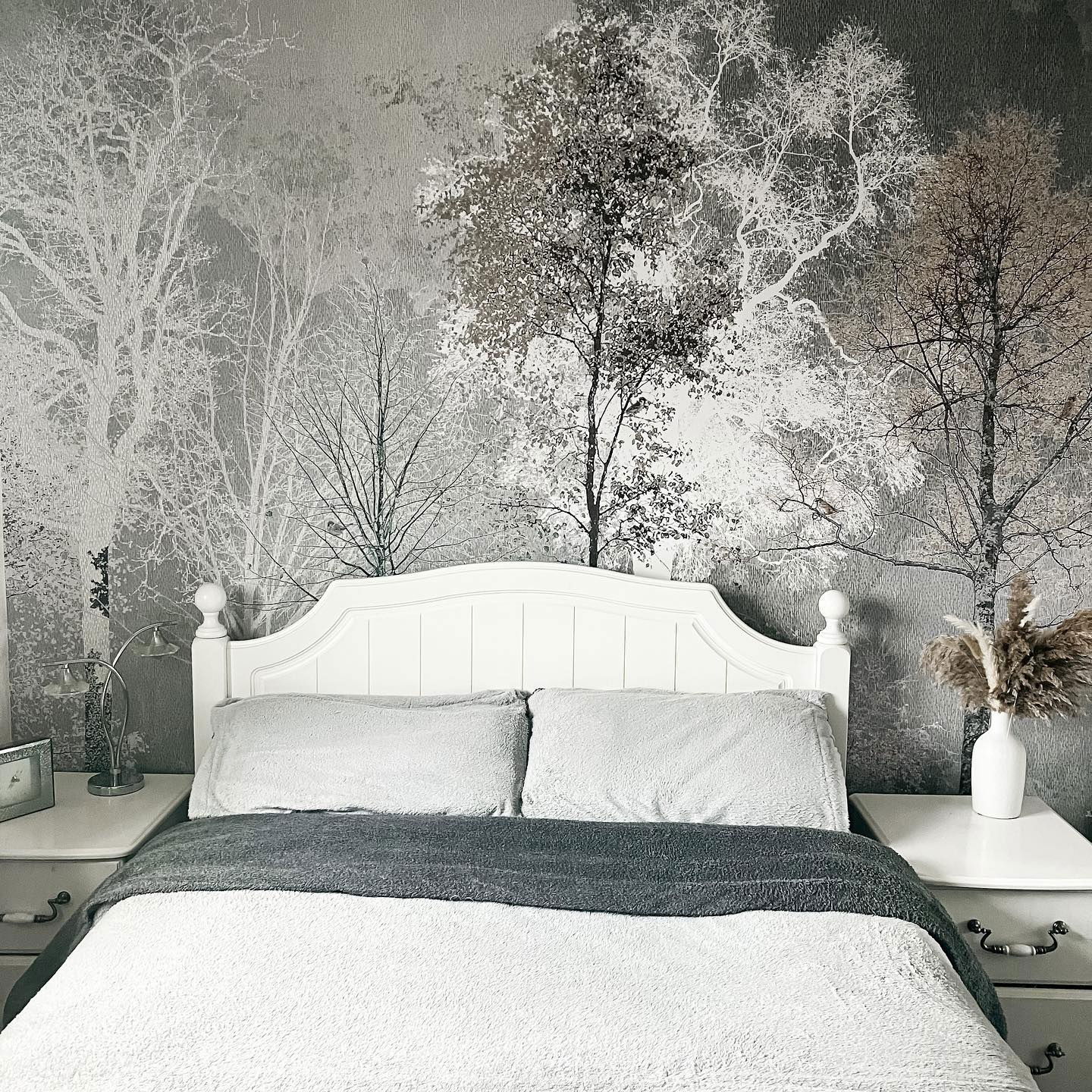 Bedroom with white bed, gray blanket, and tree-themed wallpaper. Nightstands flank the bed.