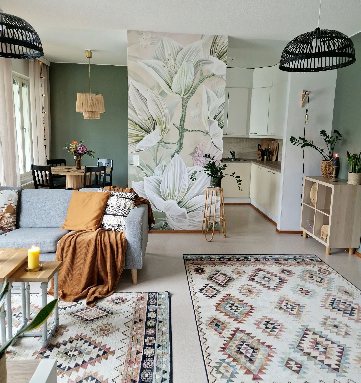 Sala de estar con mural floral, sofá gris y alfombras estampadas. Área de la cocina visible al fondo.