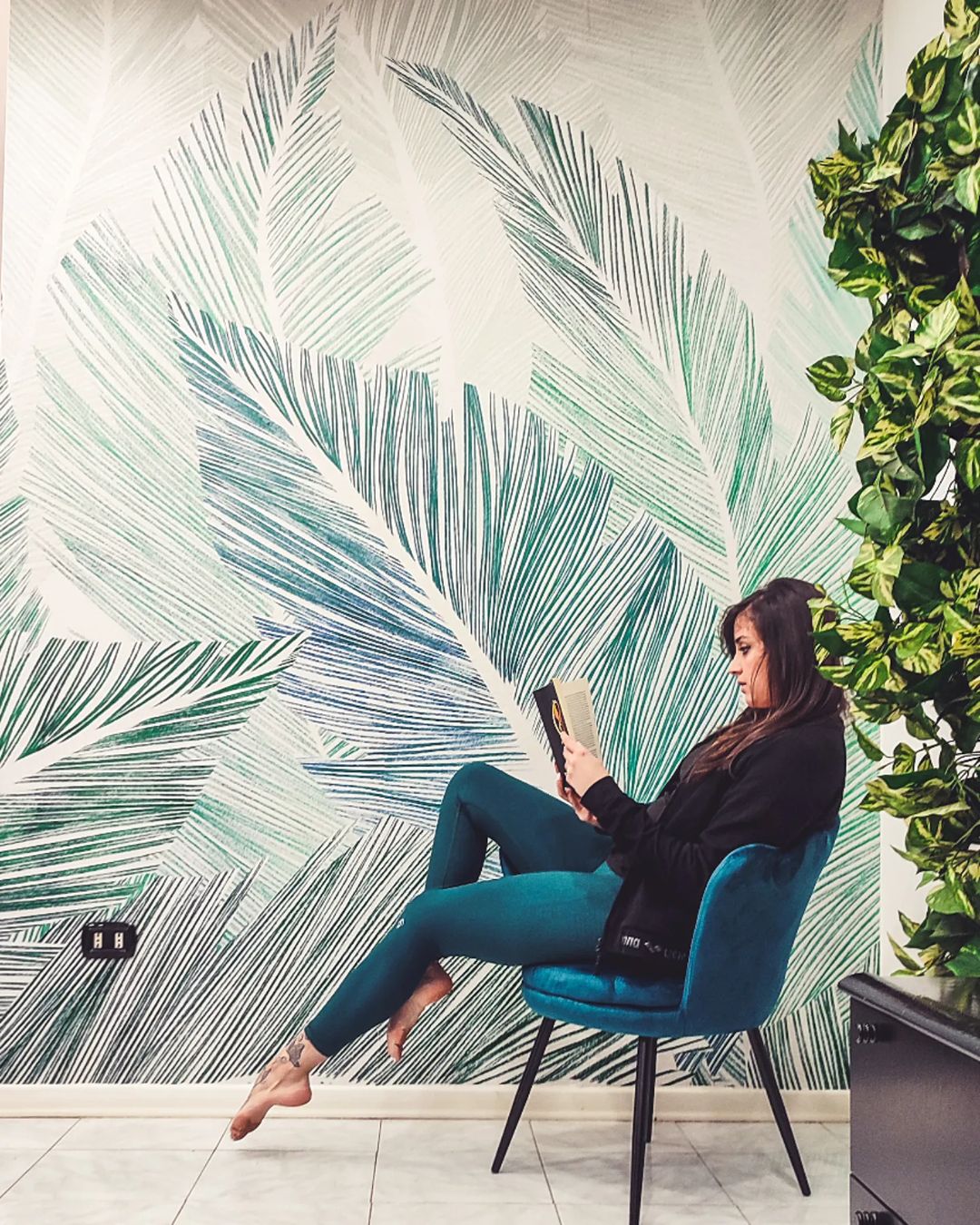 Woman reading a book in a blue chair against a leafy wallpaper.