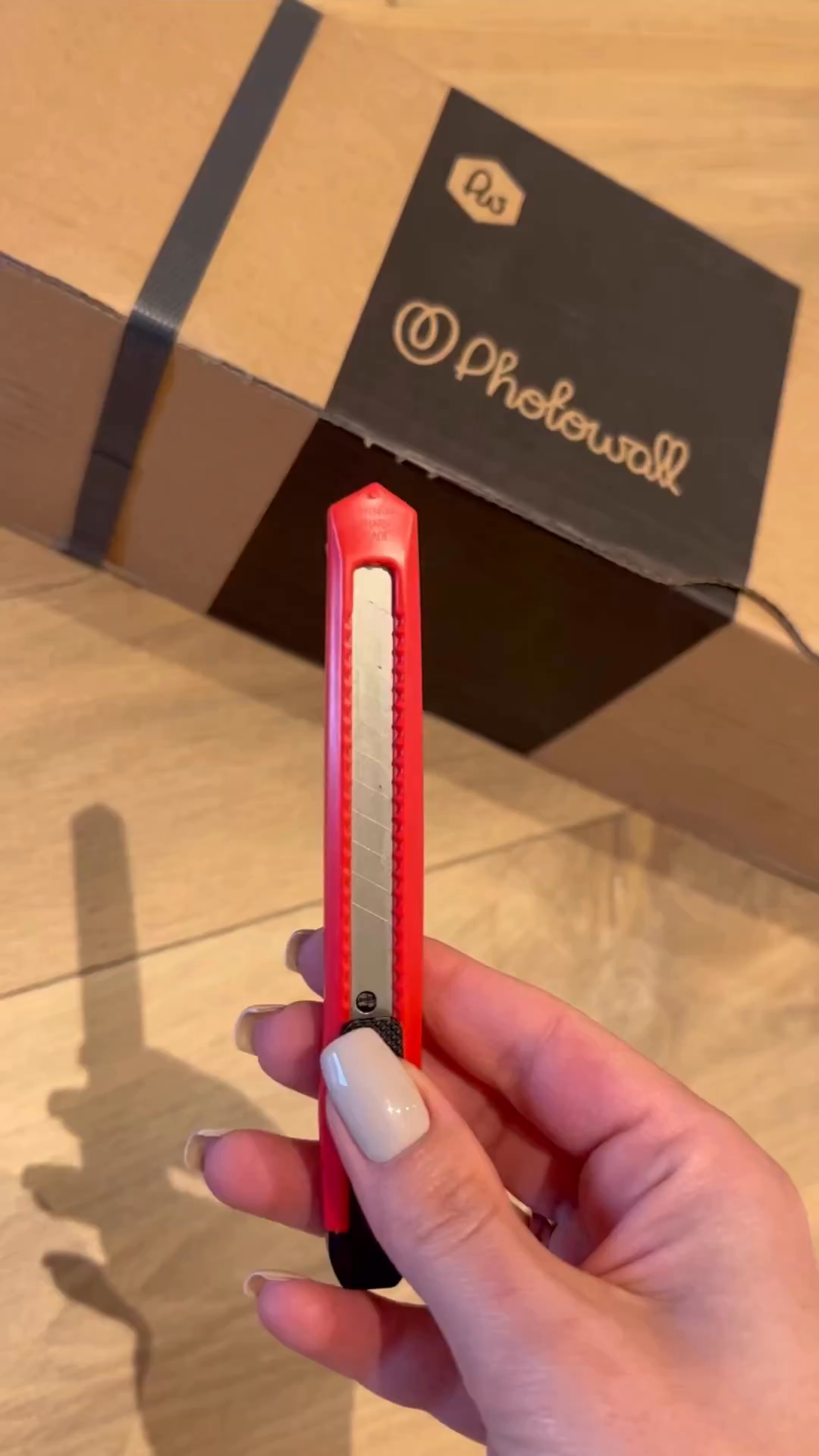 Hand holding a red utility knife in front of a Photowall package.