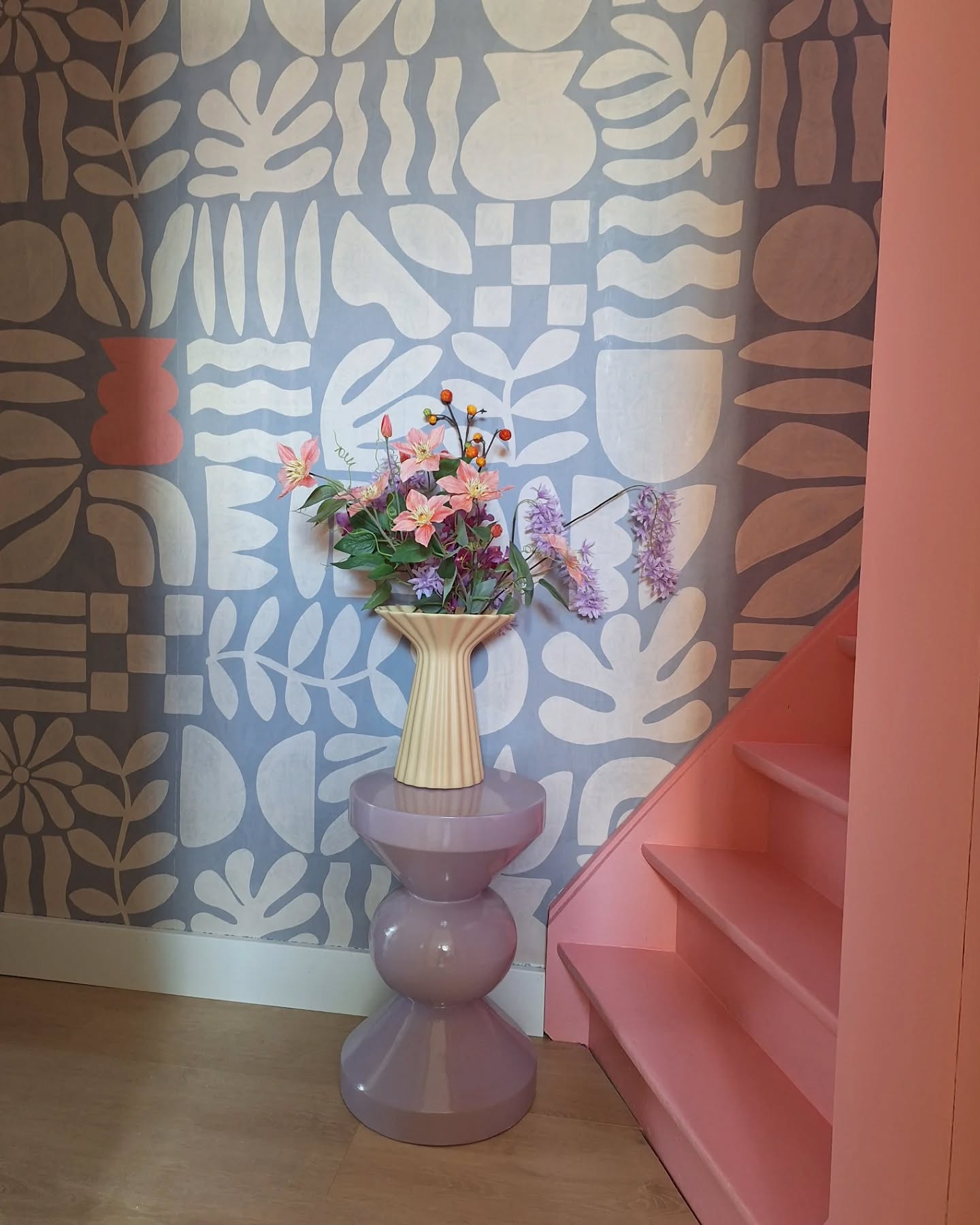 Interior with floral wallpaper, pink stairs, and a purple side table holding a vase of flowers.