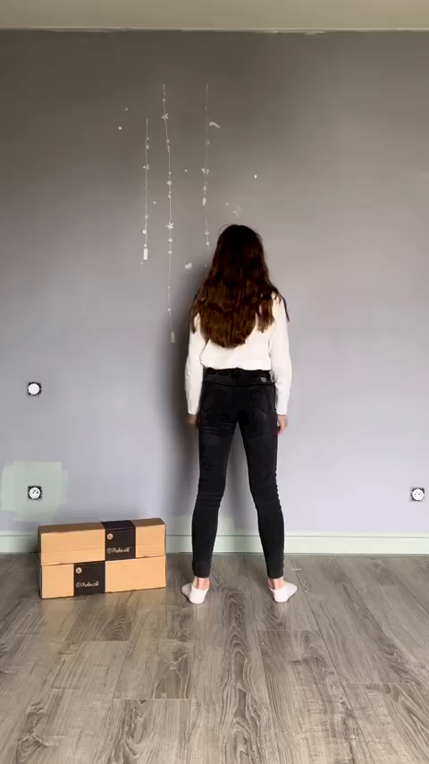 A woman stands in black jeans and a white sweater in front of a gray wall. Two boxes with the "Rebell" logo are on the floor.