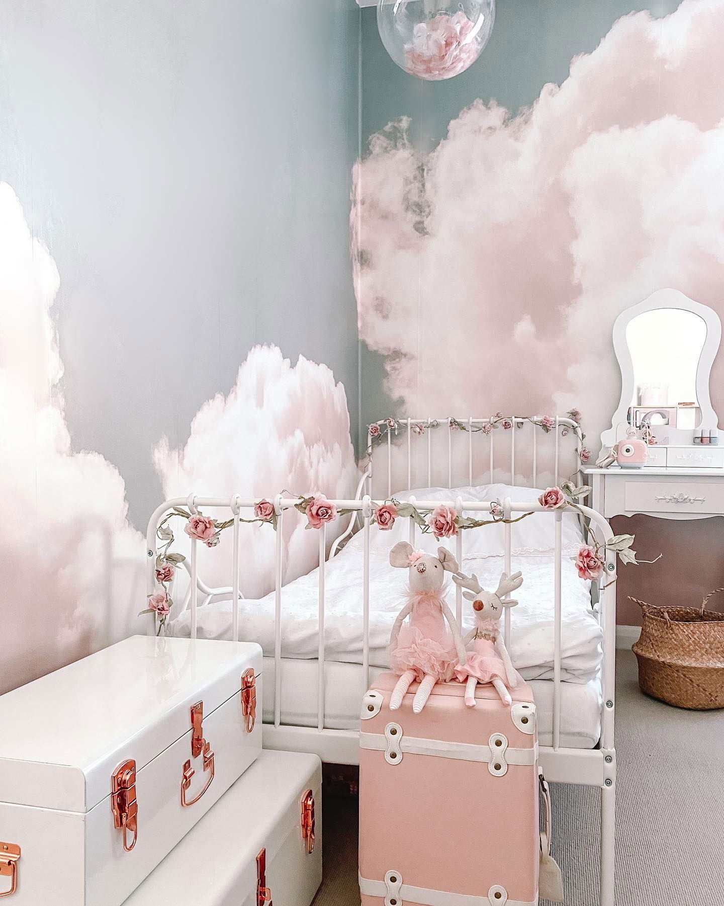 Whimsical girl's room with cloud wallpaper, white metal bed with rose garland, and pink trunk with stuffed animals.