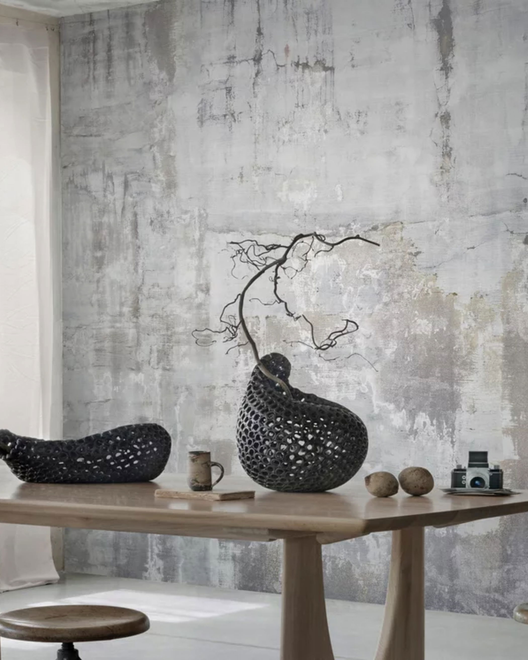 Interior with wood table, stool, and textured gray wall. Table has black woven vases, mug, and camera.