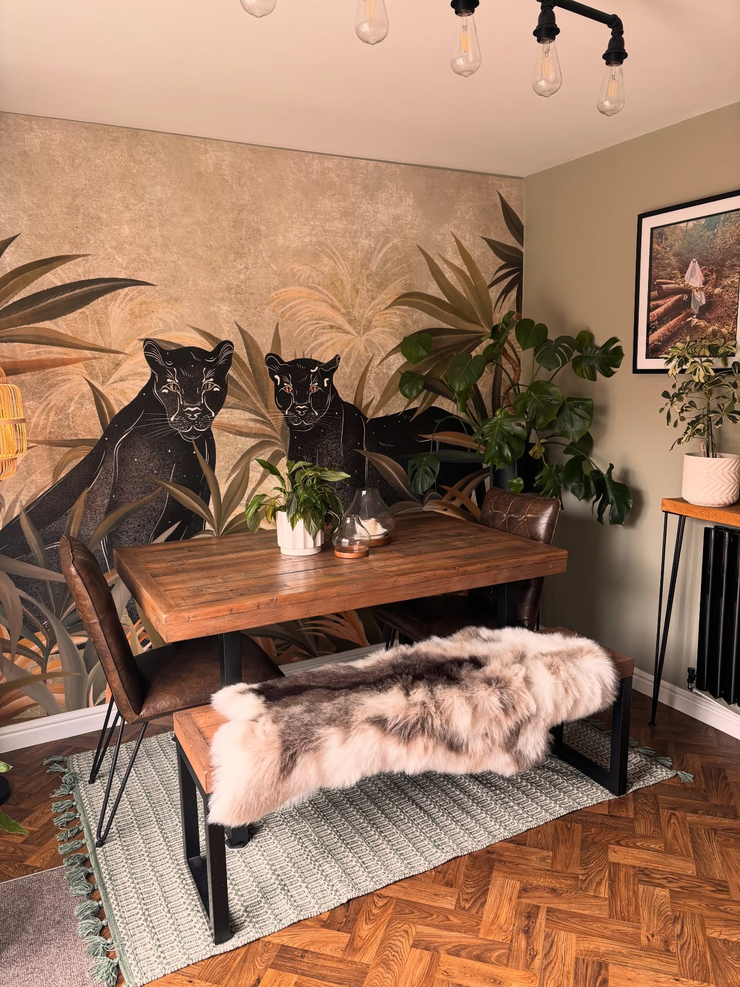 Tropical-themed dining area with a mural of two black panthers and green foliage. A rustic wooden table and bench, covered with a fluffy brown and white throw, are paired with two brown leather chairs on a green rug.