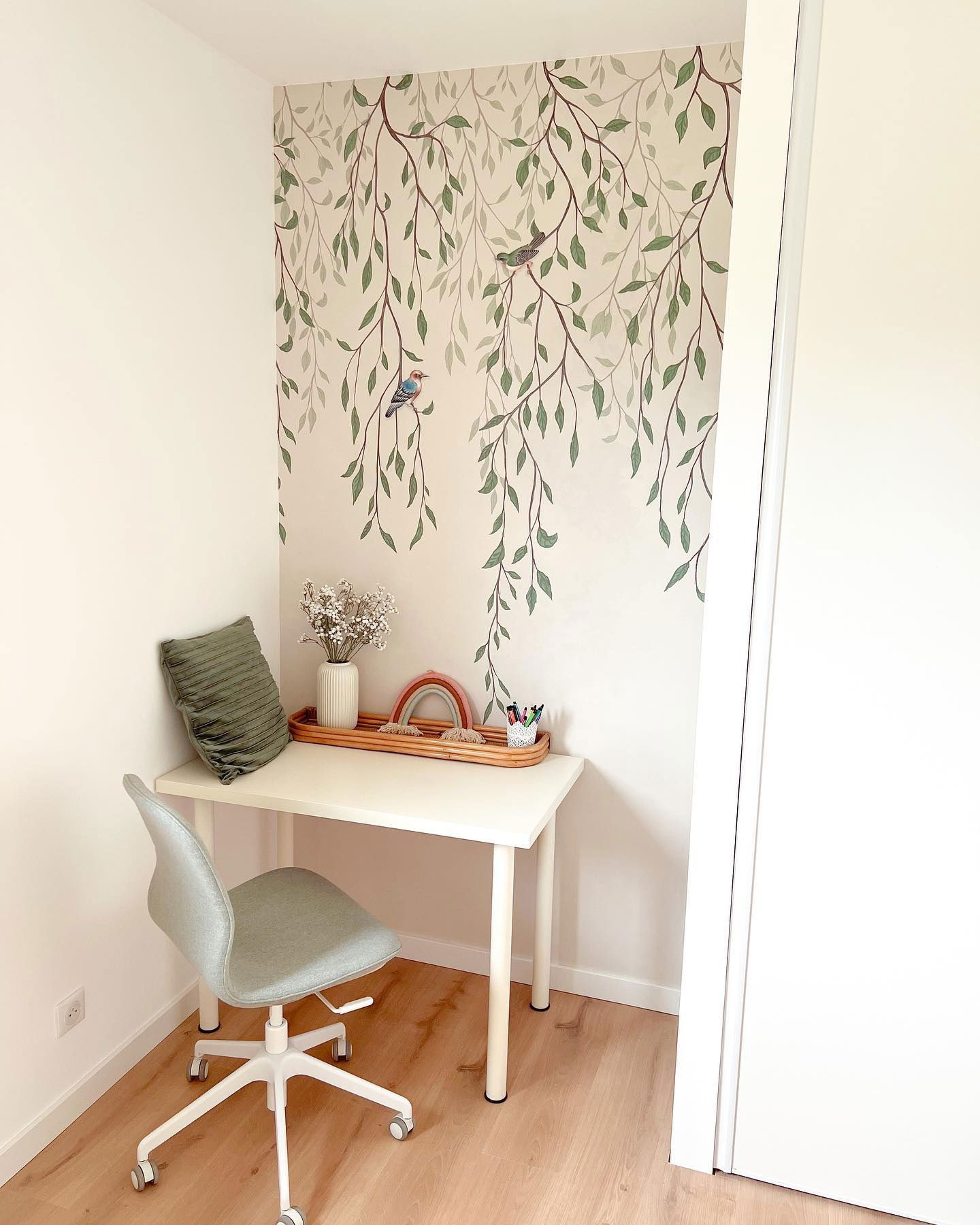 Desk with white vase, rainbow decor, and pencil holder. Wall has a leafy branch and bird design. Gray chair.