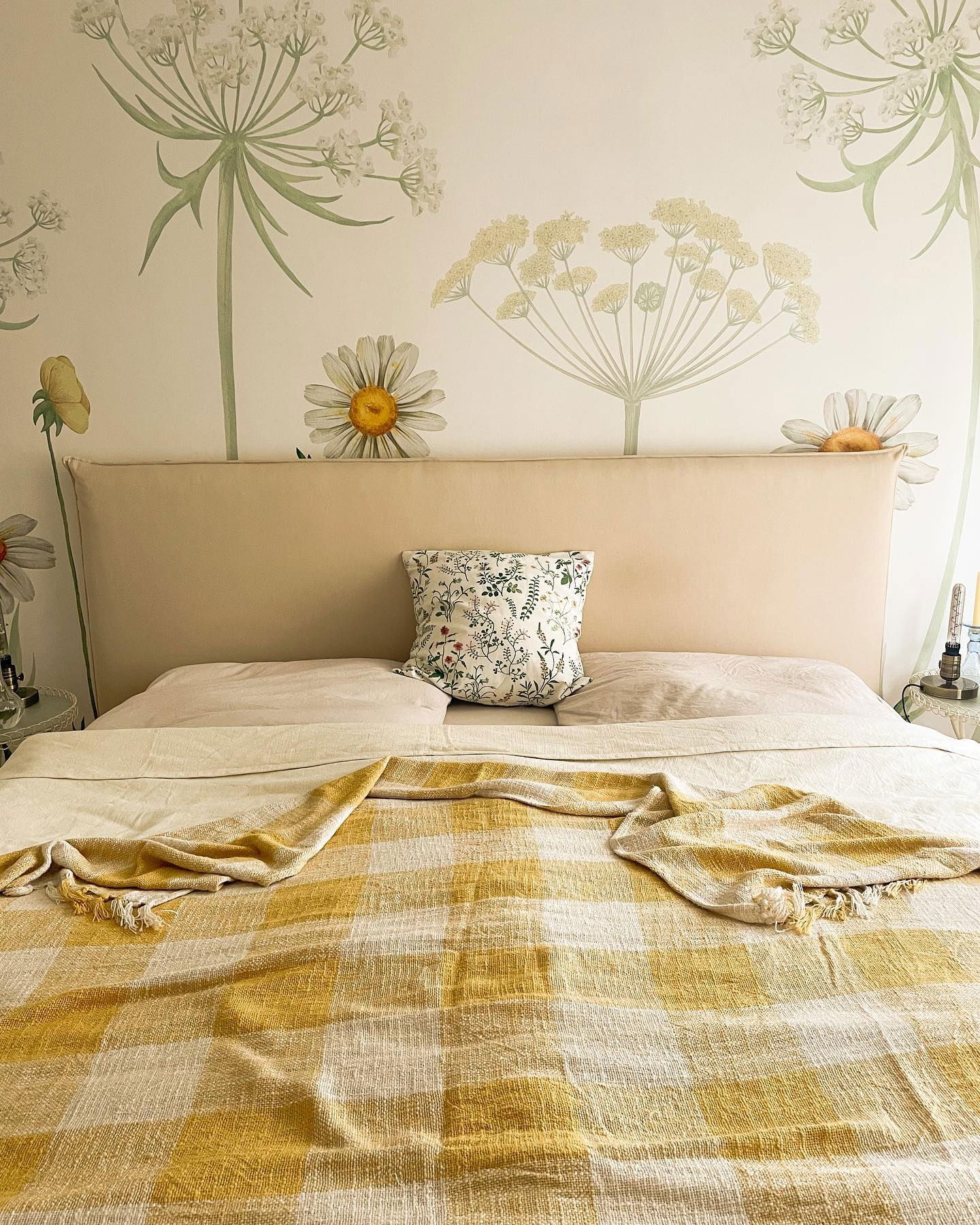 Schlafzimmer mit floraler Tapete, beigem Kopfteil und gelber karierter Überwurfdecke.
