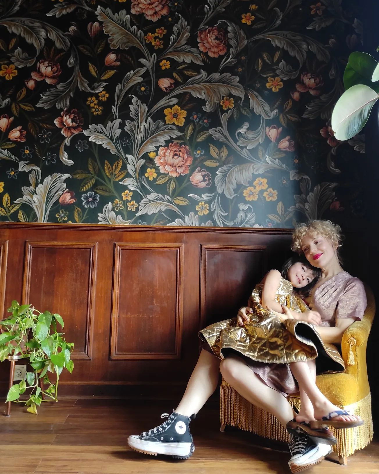 A woman and child embrace in a yellow chair against a floral wallpaper backdrop. The woman wears Converse sneakers.