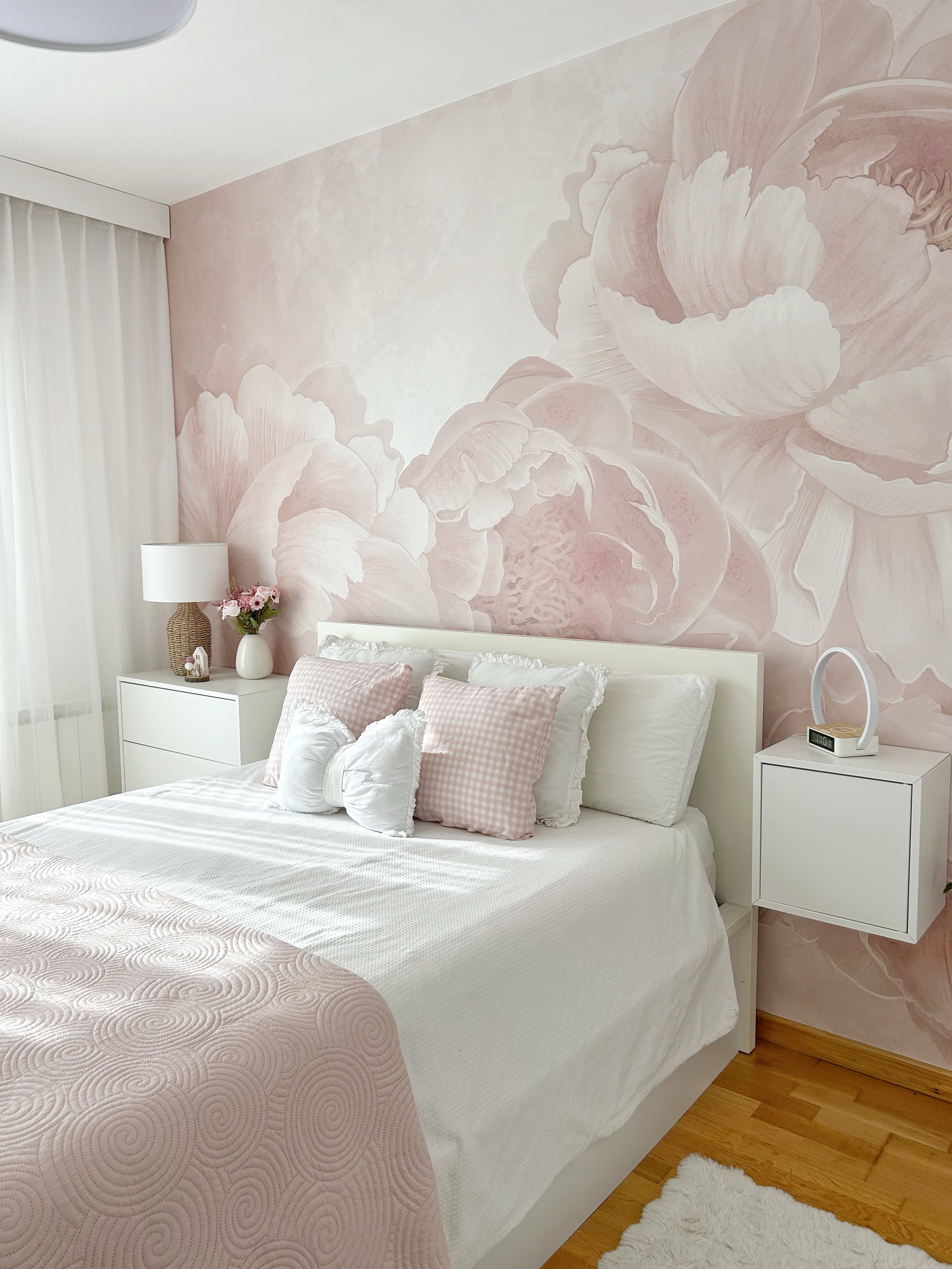 Una camera da letto con un letto bianco, cuscini decorativi rosa e una coperta a motivi rosa. La parete ha una carta da parati con grandi peonie rosa. Comodini bianchi su ciascun lato.