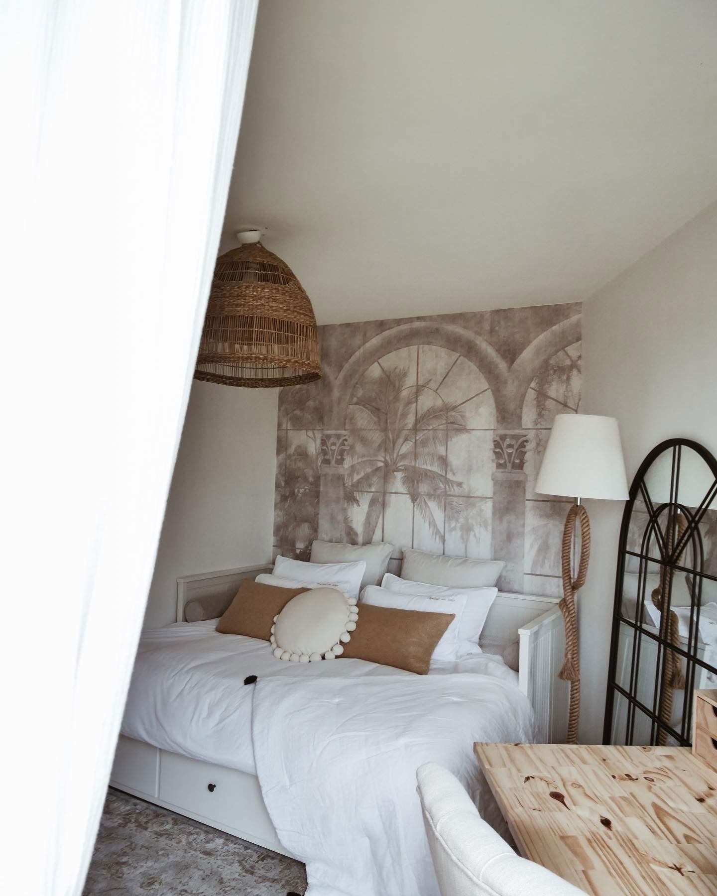 Cozy bedroom with white bedding, brown pillows, woven pendant light, and palm tree wallpaper.