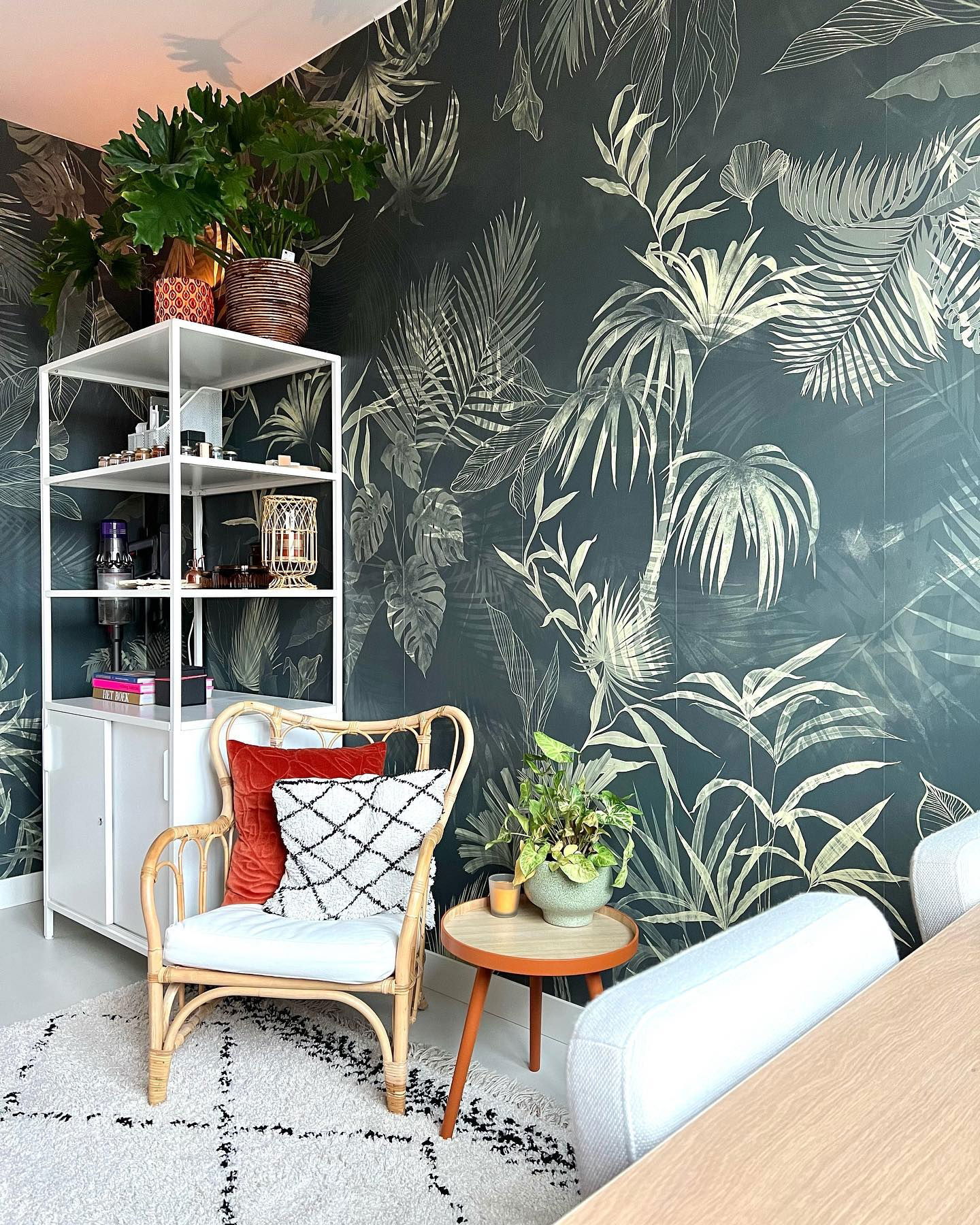 Eclectische woonkamer met tropisch behang, rotan stoel, witte stellingkast en een vloerkleed met motief.