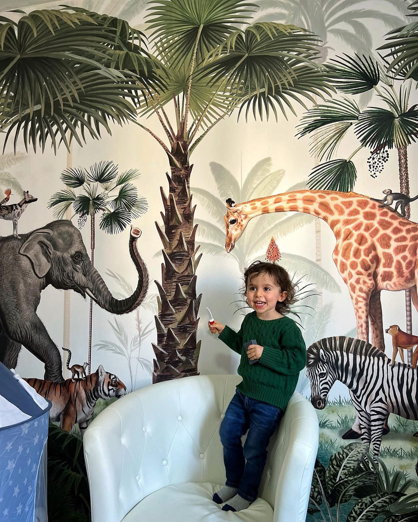 Kind in groene trui en spijkerbroek staat in een witte stoel tegen een jungle dierenbehang.