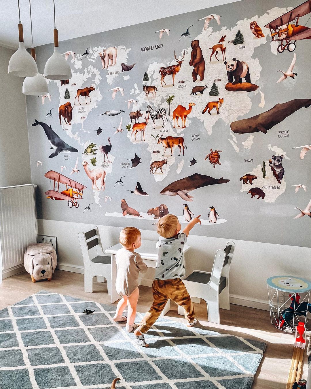 Two toddlers in a playroom with a world map wall decal featuring animals, a blue patterned rug, and white furniture.