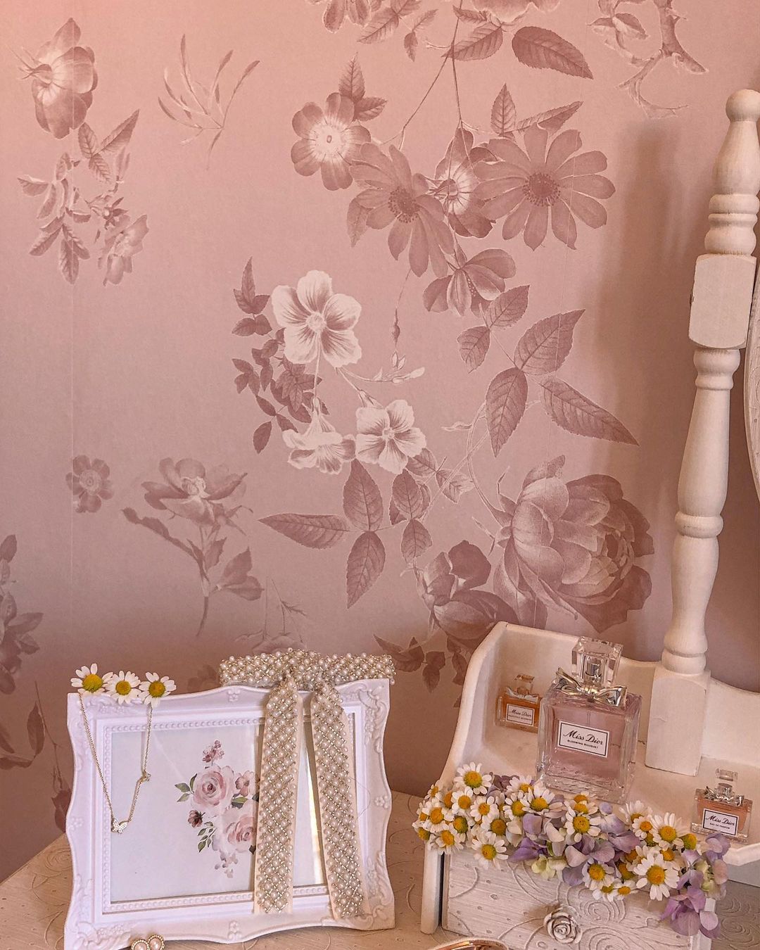 Bloemig roze behang met een witte wastafel met Miss Dior-parfum, madeliefjes en een parelmoerlijst.