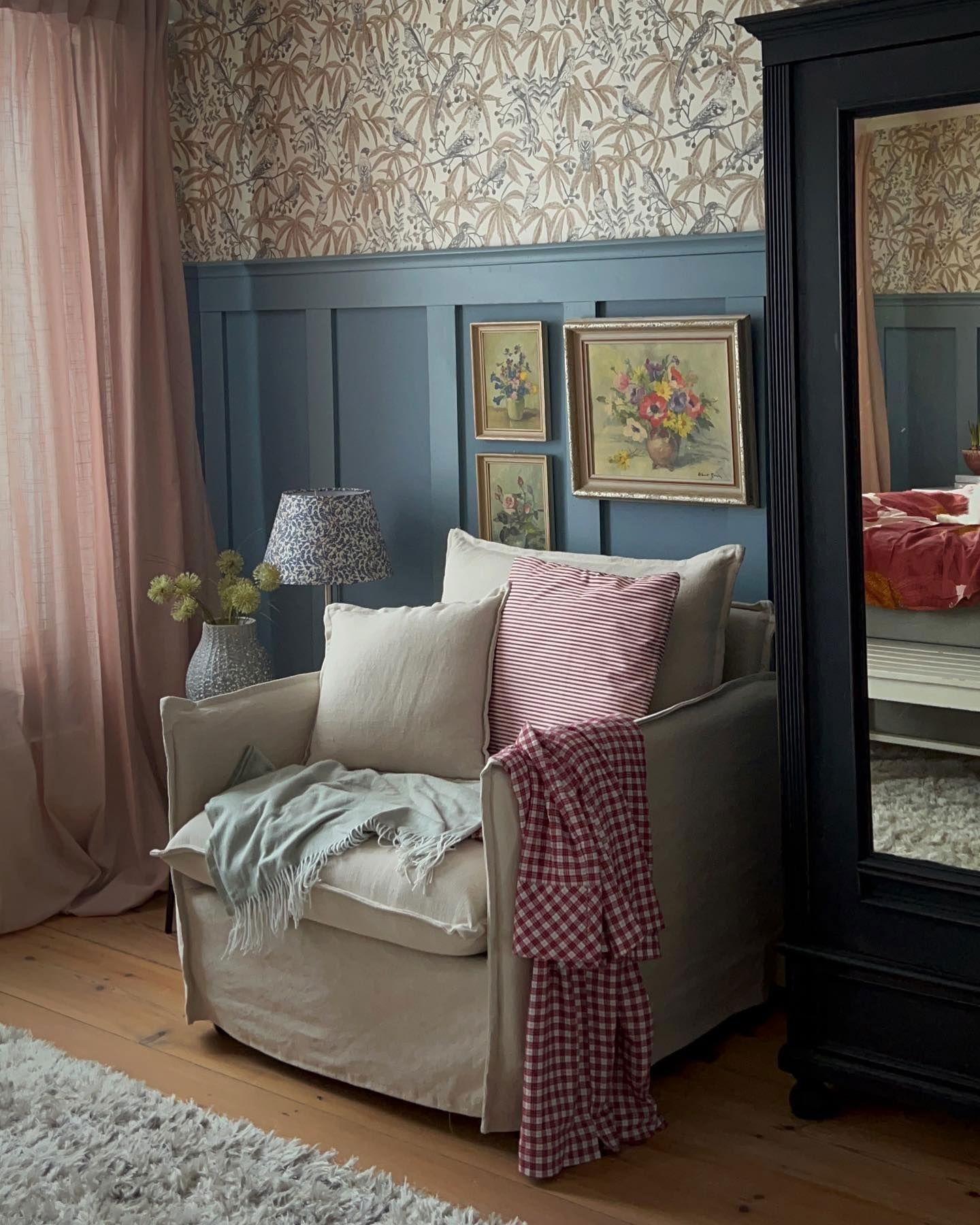 Interior acogedor de una habitación con sillón, pared con paneles azules y papel tapiz floral. Mantas y almohadas adornan la silla.