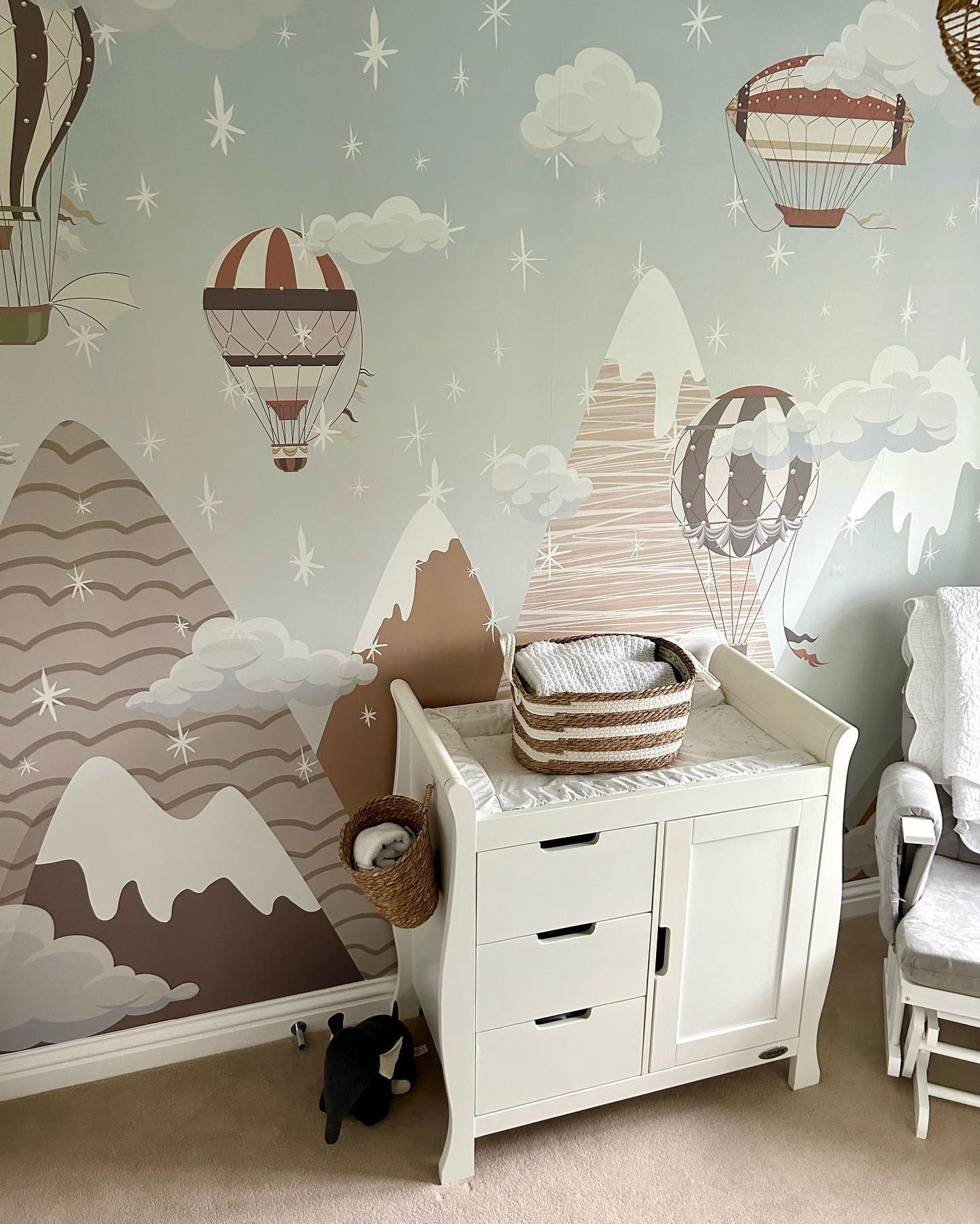 Kinderzimmer mit Berg- und Heißluftballon-Tapete, weißer Wickeltisch und geflochtenen Körben.