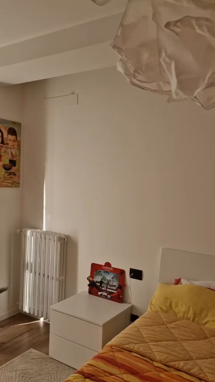 Bedroom with white walls, radiator, nightstand with Miraculous Ladybug toy, and bed with yellow and orange striped comforter.