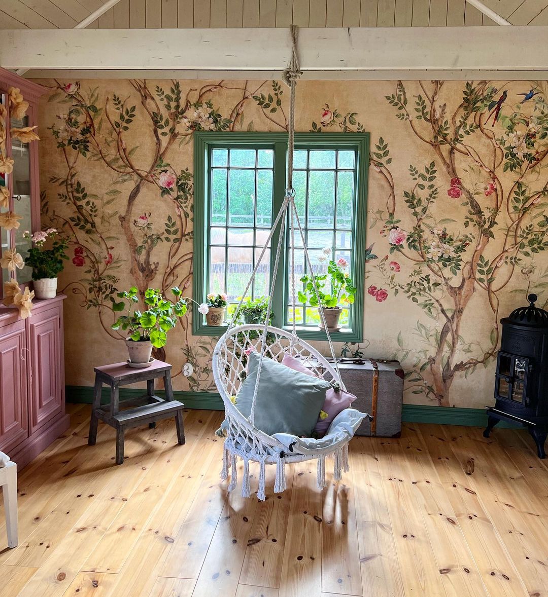Hyggeligt værelse med blomstret tapet, grønt vindue og hængende macramé-stol med puder.