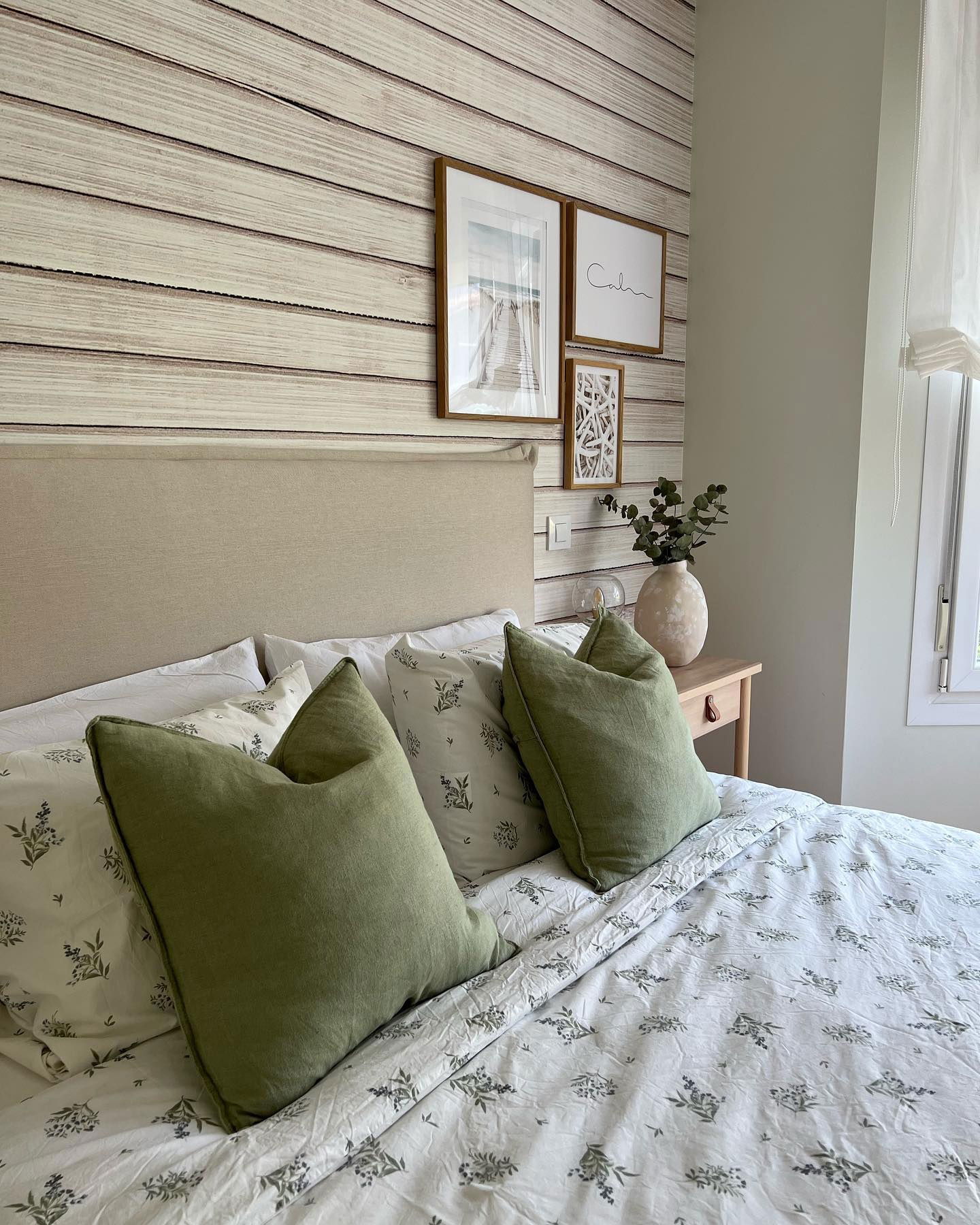 Bedroom with floral bedding, green pillows, and wood-paneled wallpaper. Framed art hangs above a beige headboard.