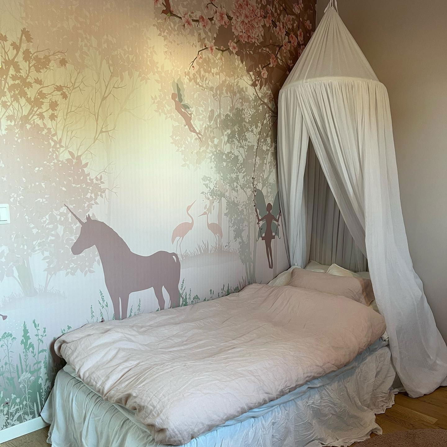 Bedroom with pink duvet, white canopy, and whimsical wallpaper featuring unicorns, fairies, and trees.