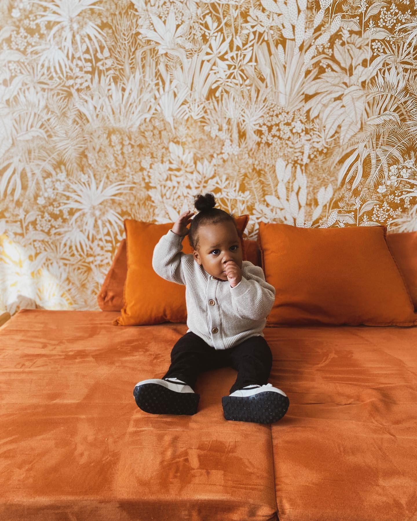 A baby sits on an orange bed with matching pillows, wearing a cardigan and black pants.