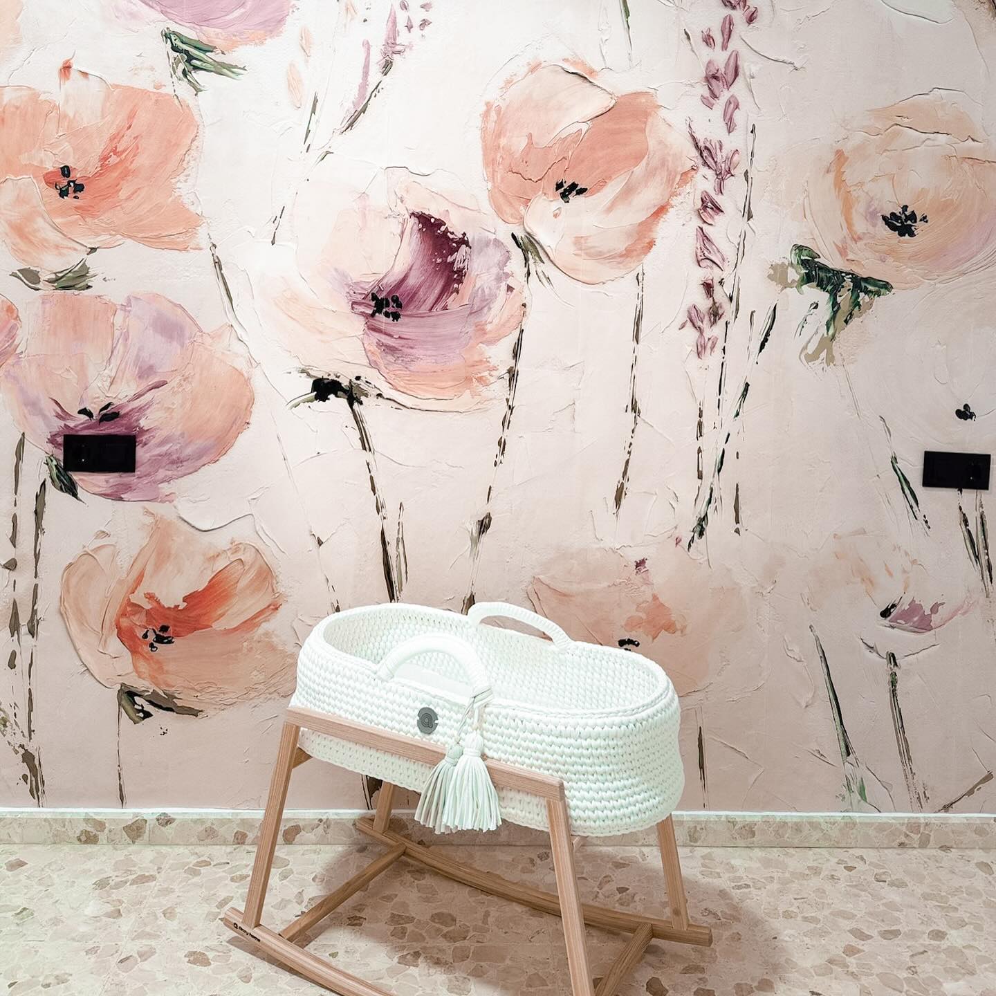 Weißer gewebter Baby-Stubenwagen mit Holzständer, Quastendetail. Florale Tapete im Hintergrund.