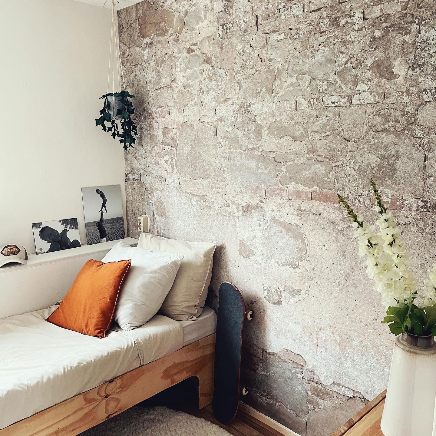 Schlafzimmer mit freiliegender Ziegelwand, Bett mit orangefarbenem Kissen, Skateboard und Hängepflanze.
