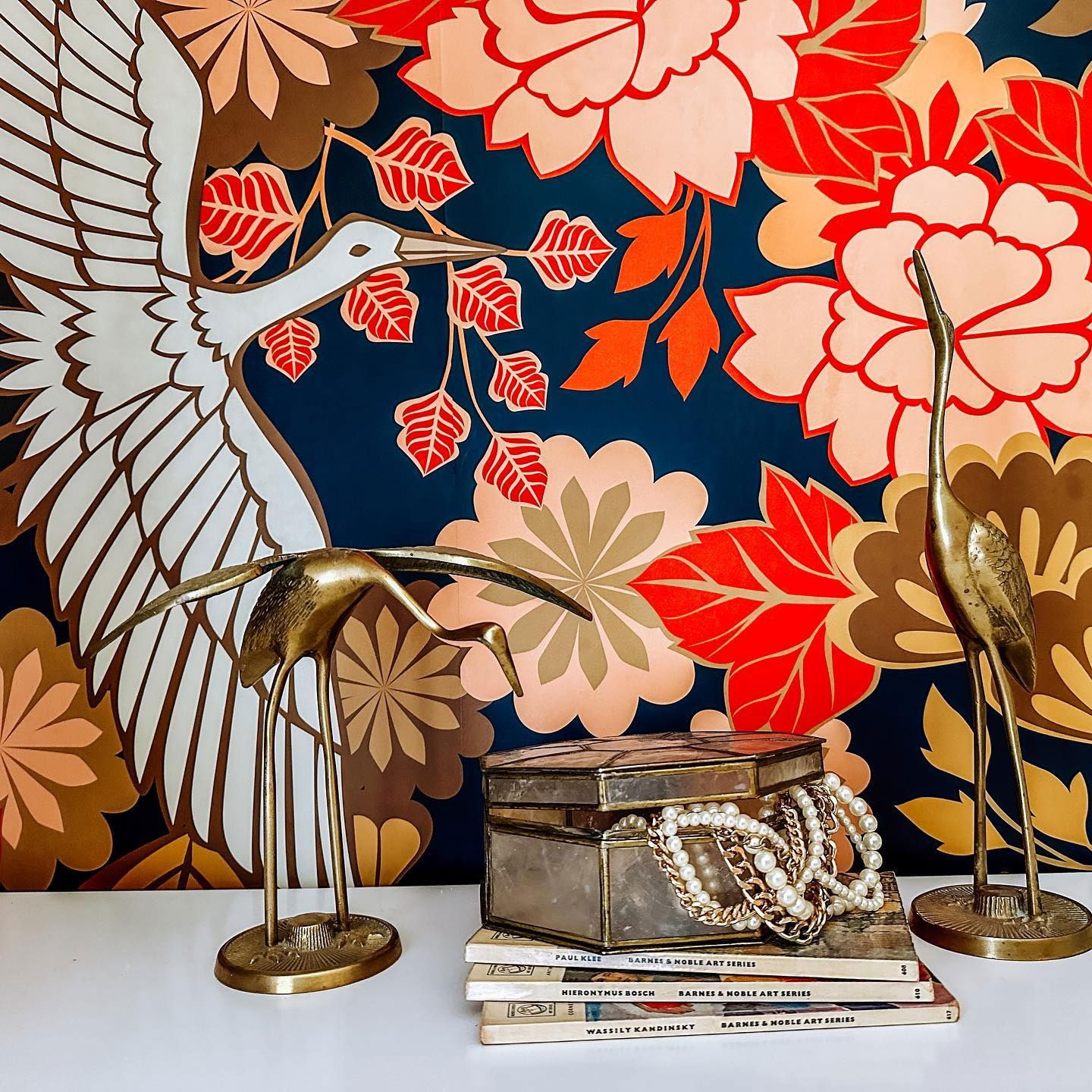 Still life with brass crane figurines, jewelry box, and art books against a floral crane print backdrop.