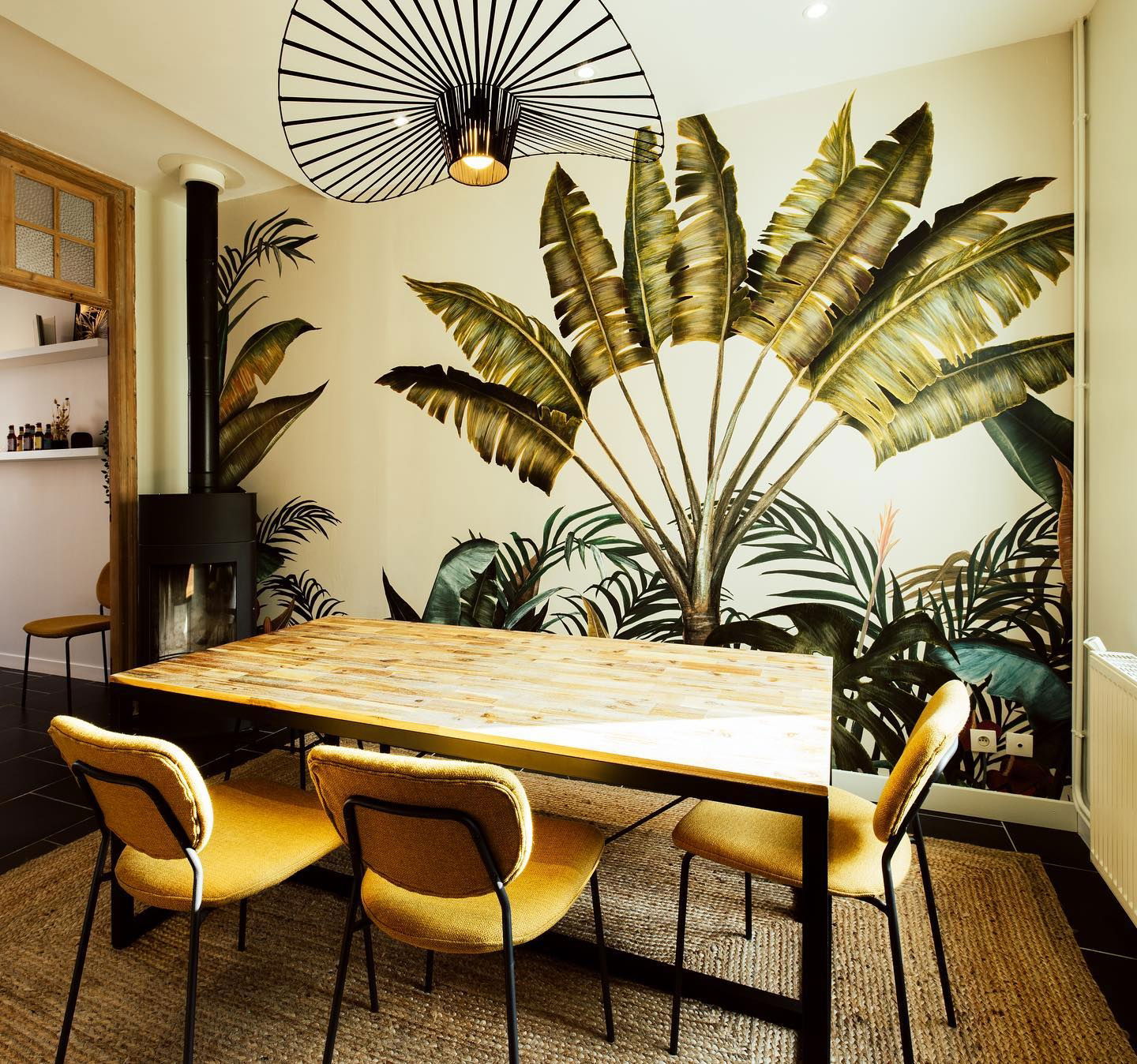 Dining room with tropical wallpaper, wood table, yellow chairs, and black metal fireplace.
