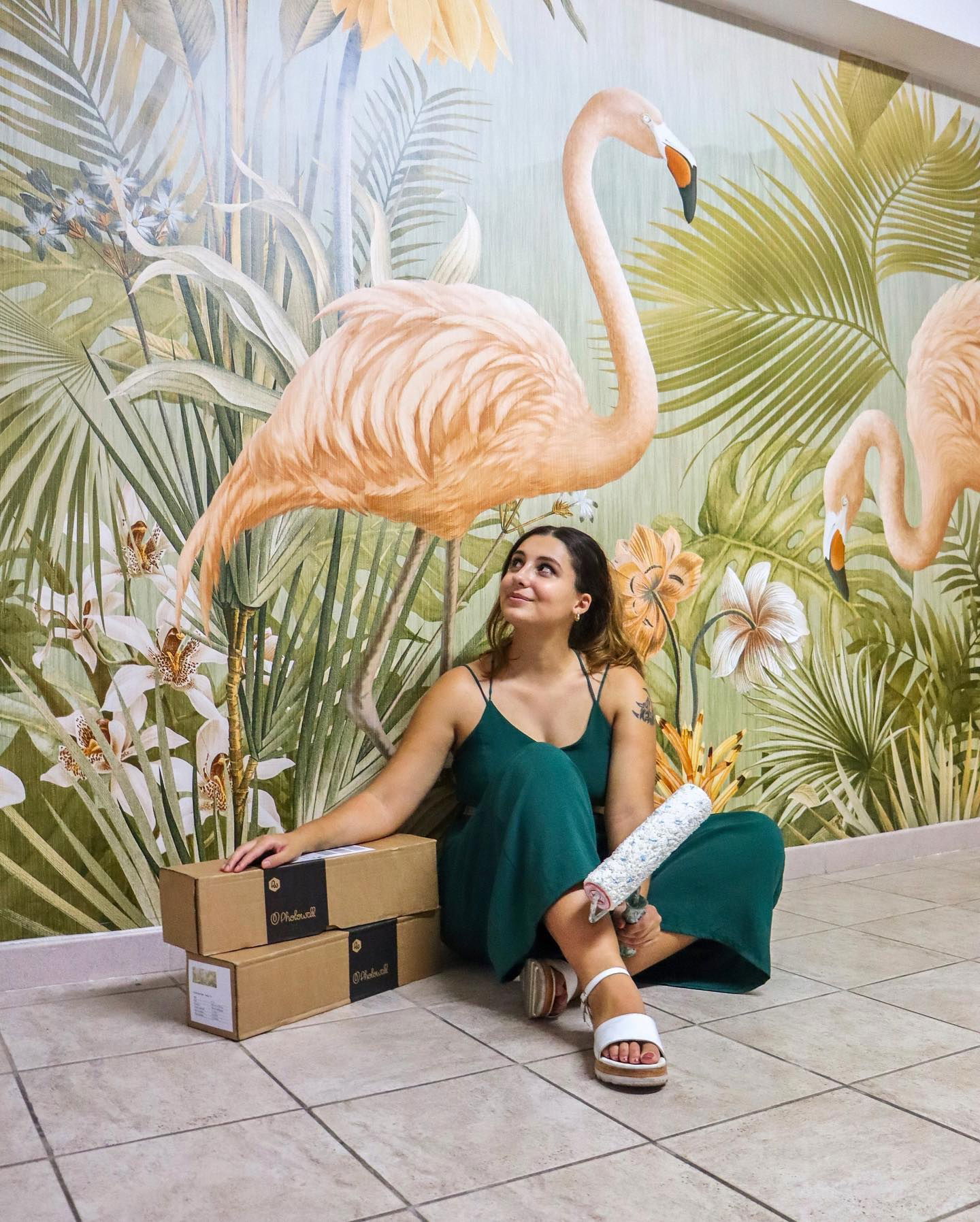 Mujer con cajas de murales Photowall y rodillo de pintura, sentada frente a un papel pintado de flamencos.