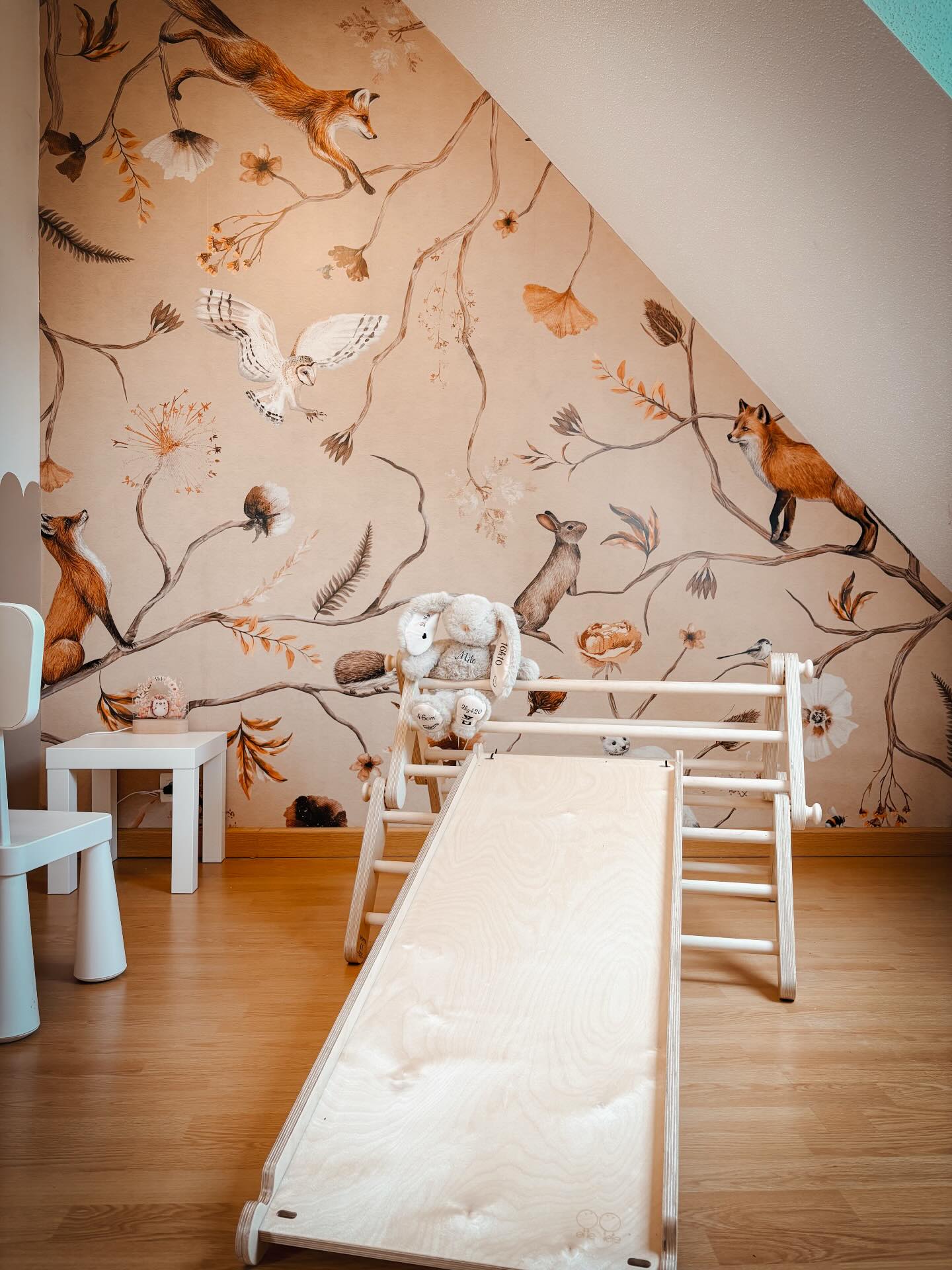 Hölzerner Pikler-Dreieck-Kletterer mit Rutsche und ausgestopftem Kaninchen in einem Kinderzimmer mit Tapete mit Waldtieren.