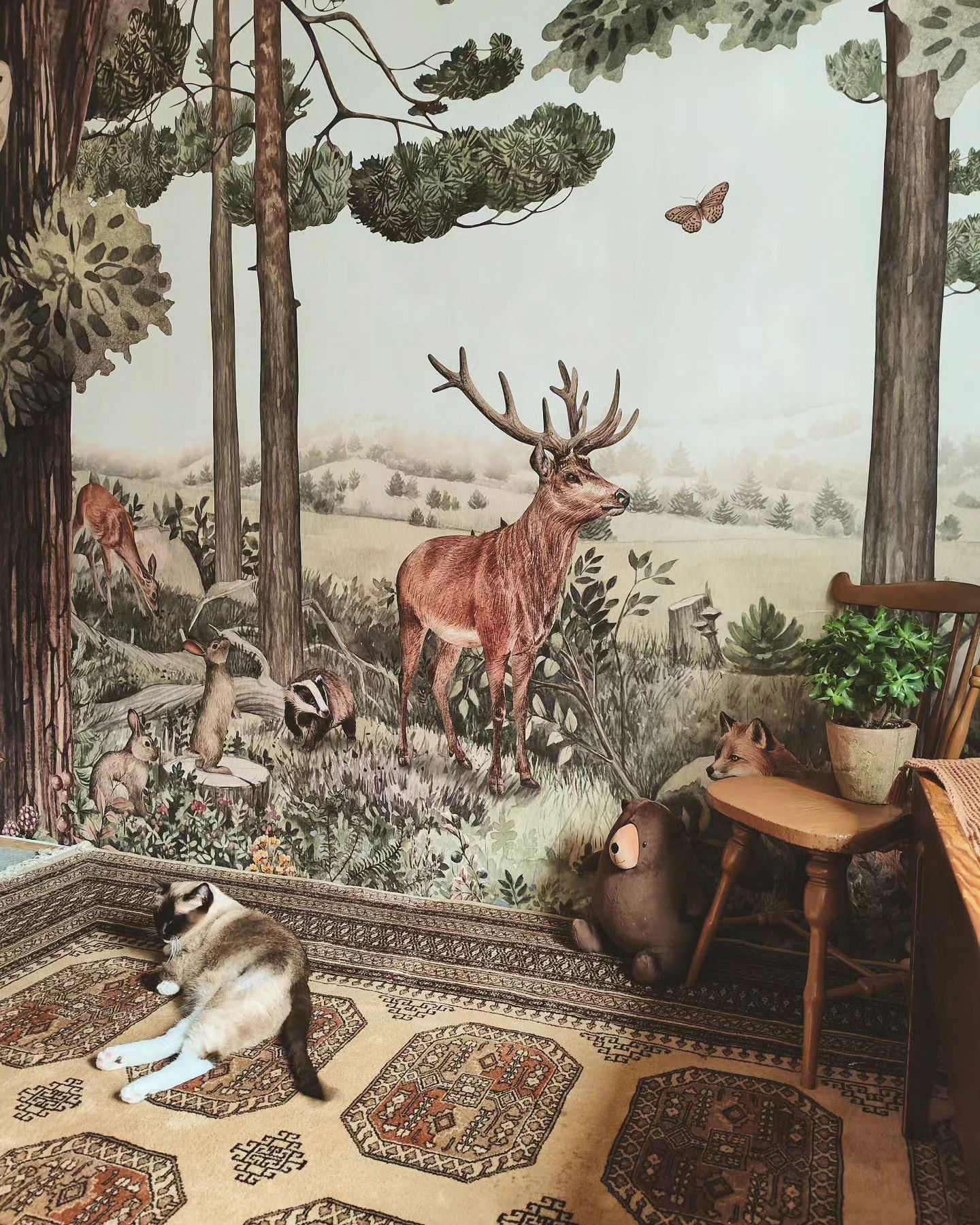 A Siamese cat lounges on a patterned rug in a room with a forest mural.