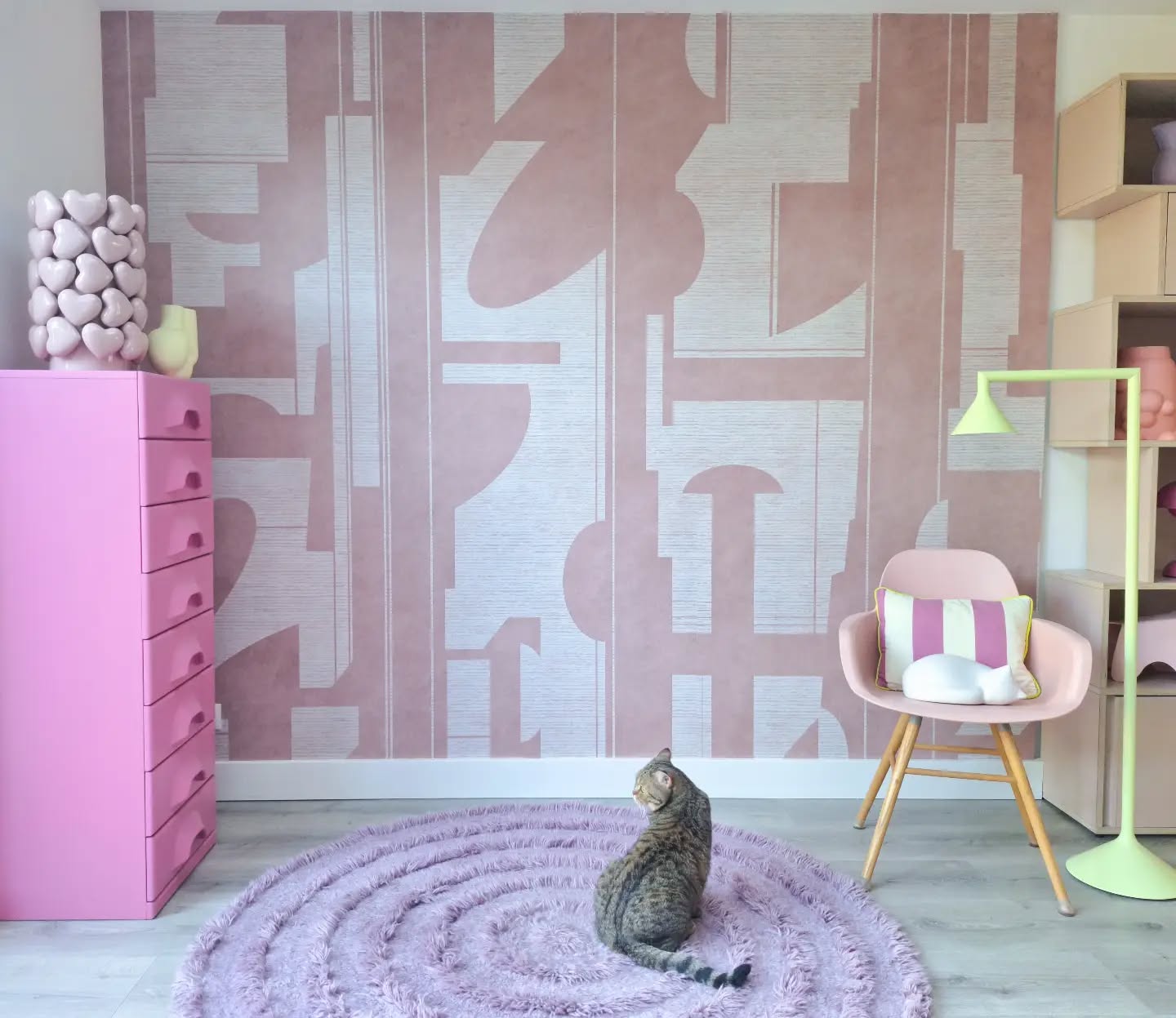 Een gevlekte kat op een rond paars hoogpolig vloerkleed. De kamer heeft roze en grijs geometrisch behang, een roze ladekast, een roze stoel en een limoengroene lamp.