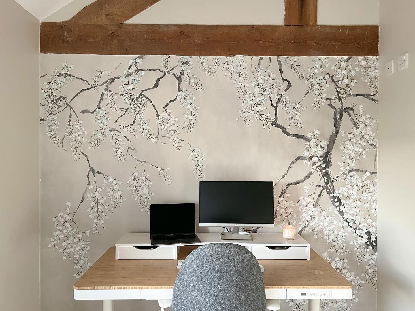 Home office setup with floral wallpaper, desk, laptop, monitor, and gray chair.