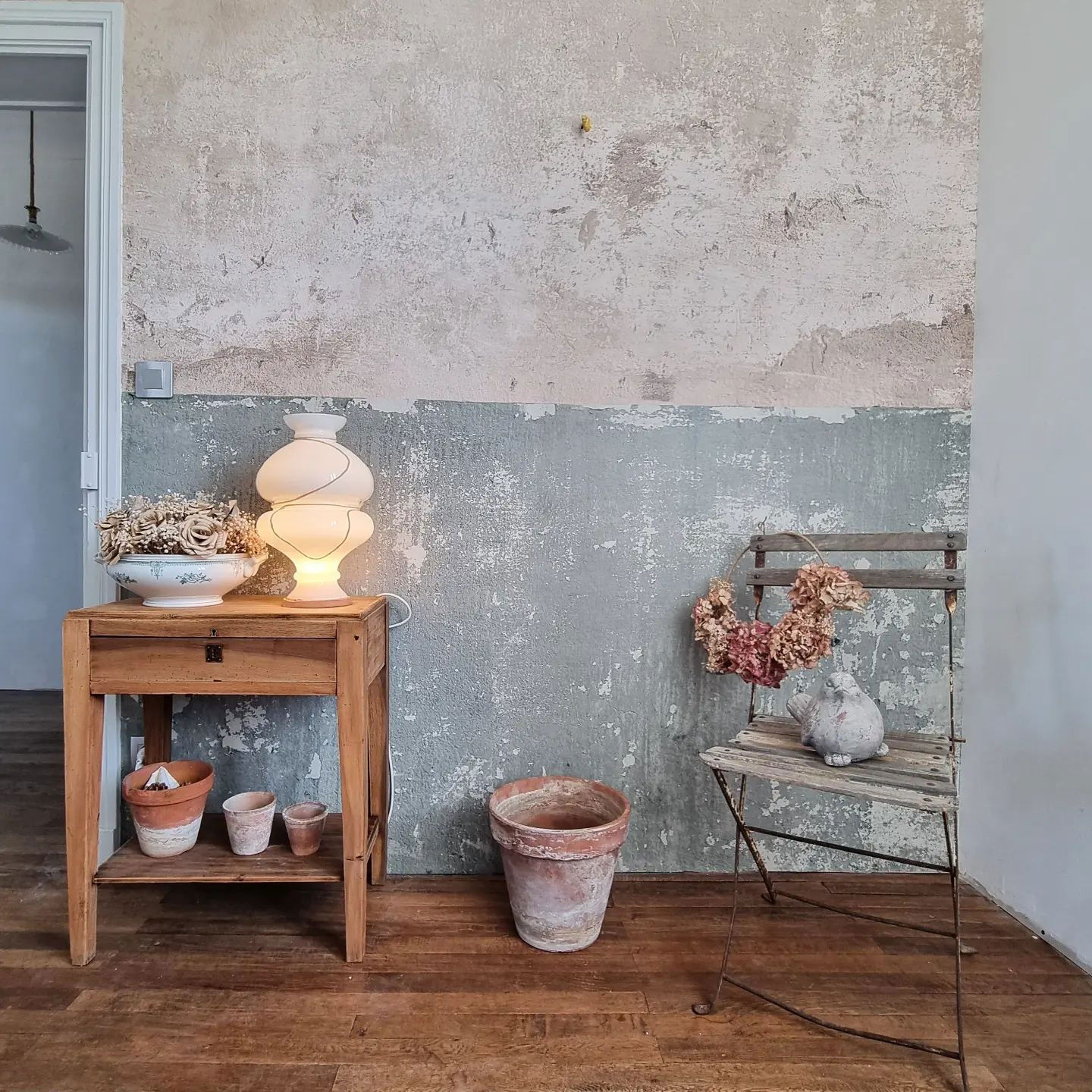 Interiör med sliten vägg, träbord med lampa och vintage-stol med krans.